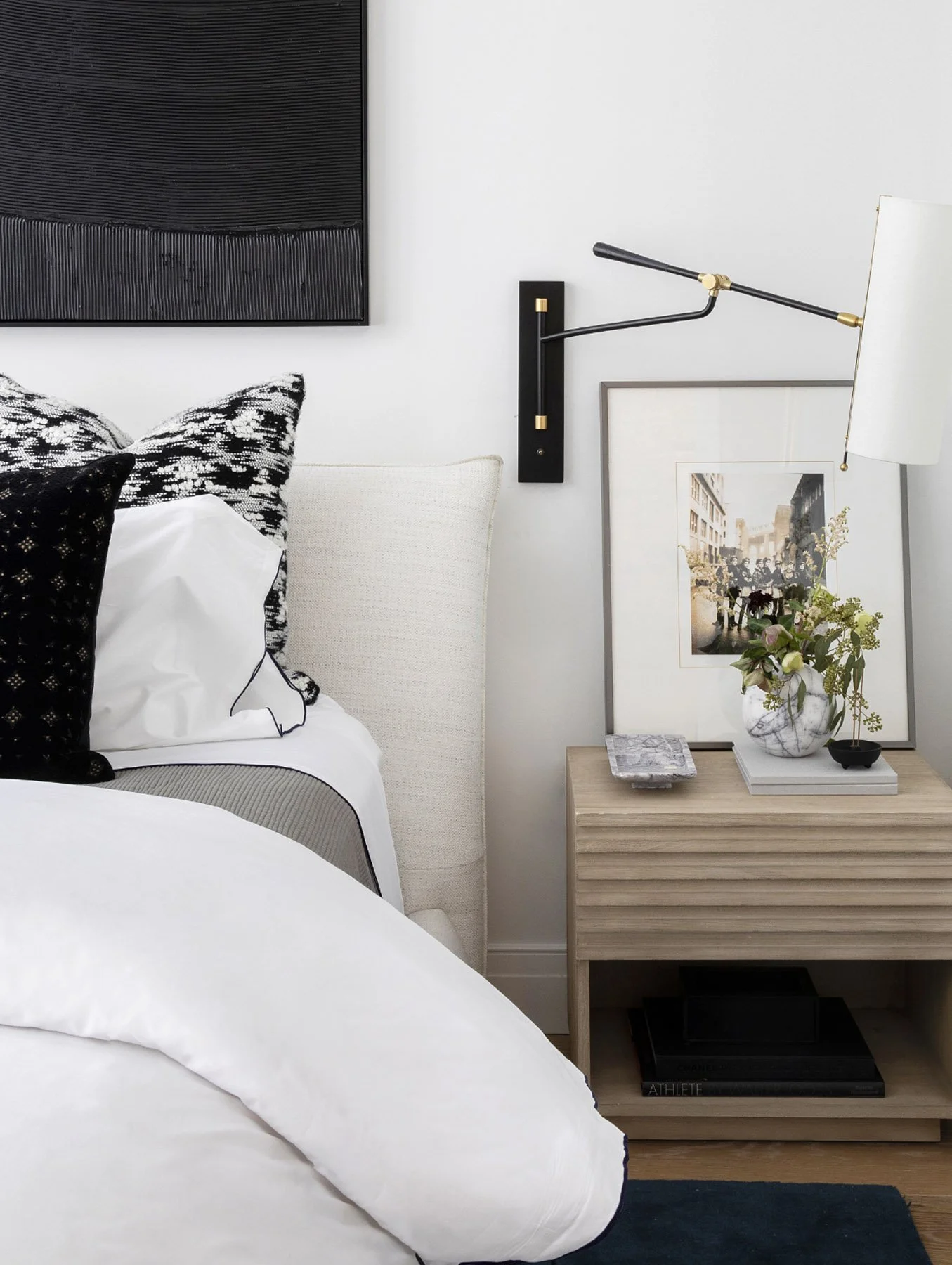 Close-up of a bed with white and black geometric pillows, nightstand with a tray, framed artwork, and decorative items, in a modern bedroom.