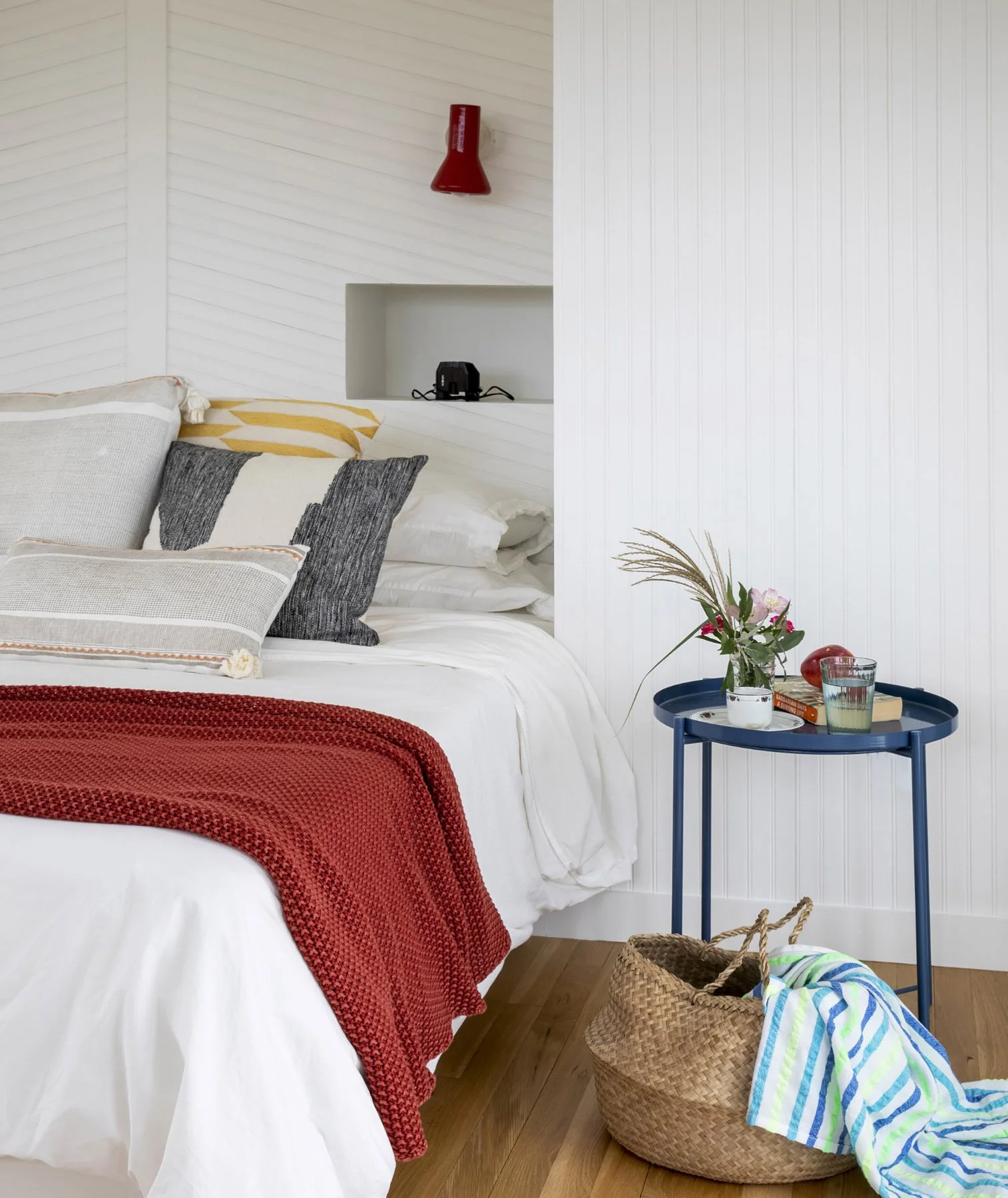 A cozy bedroom corner with a bed covered in white linens and a red knitted blanket, multiple pillows, a blue side table with a glass of water and flowers, a basket with a towel on the floor, and a built-in shelf with a camera and wall-mounted red lamp.
