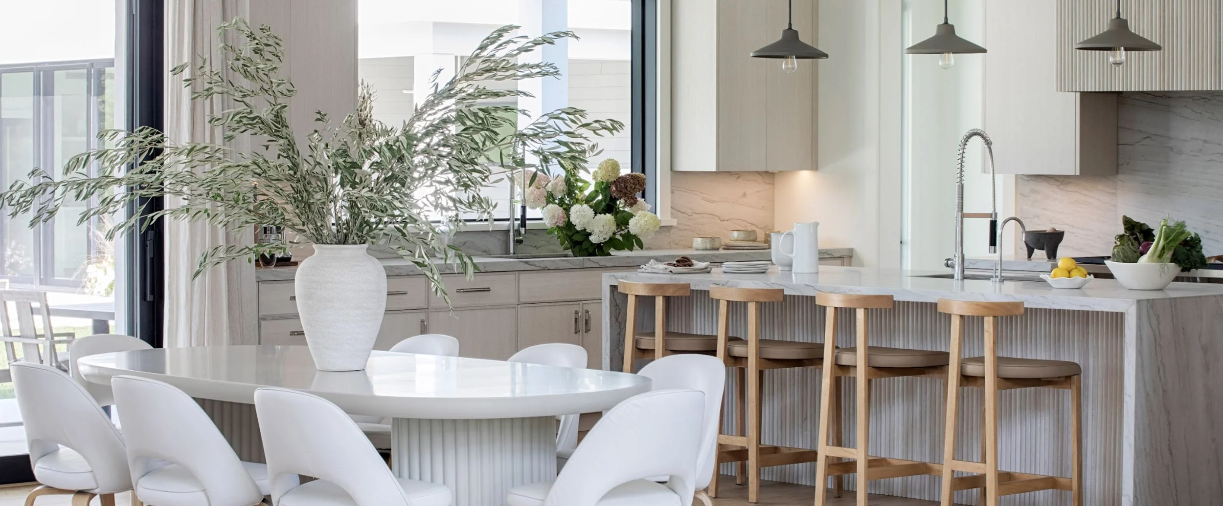 Modern kitchen with white cabinetry, marble countertops, and a kitchen island with wooden bar stools. Large window with curtains, potted plants on the counter, and a dining table with a large white vase and greenery.