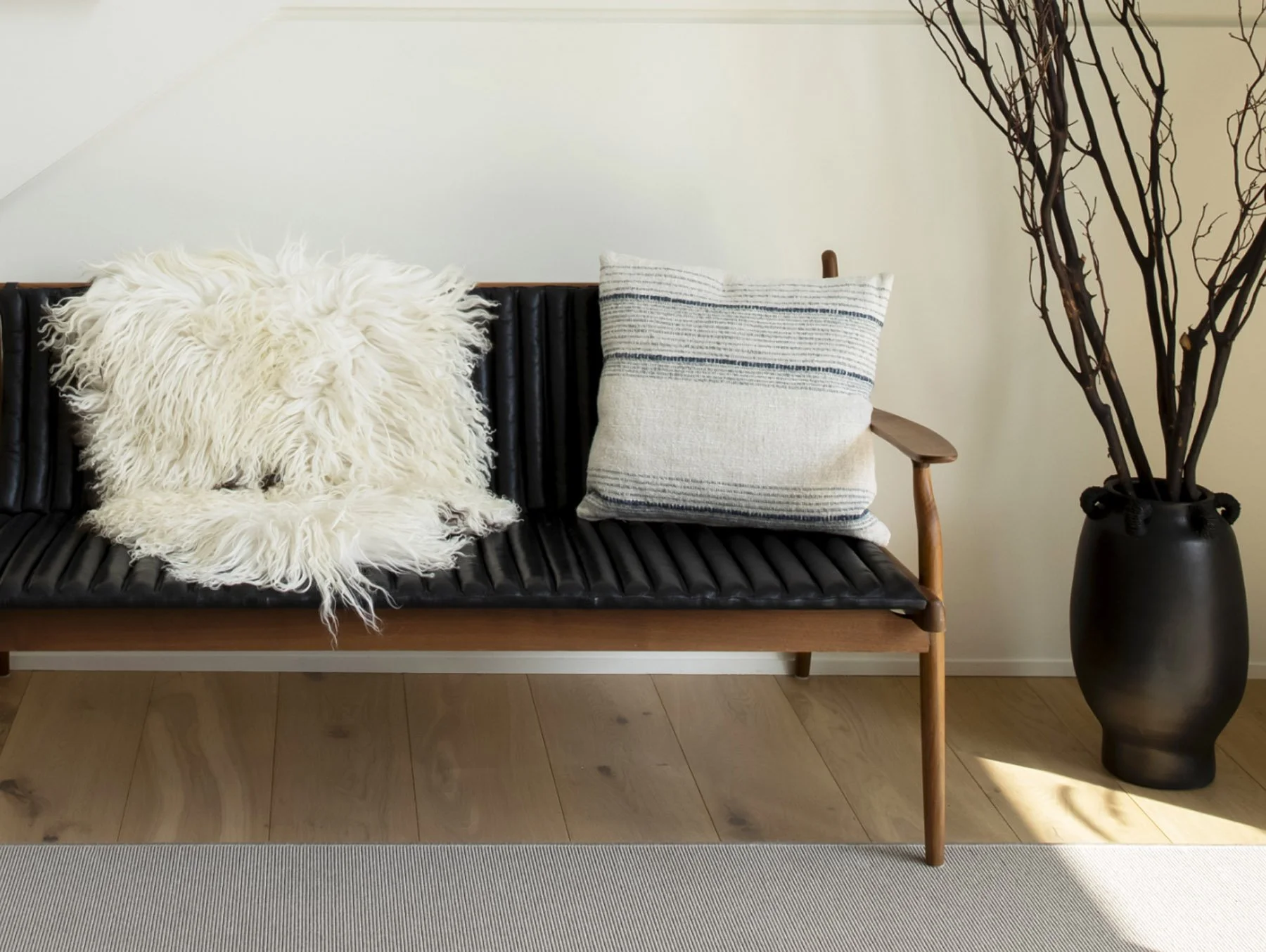 A black leather and wood bench with two pillows, one fluffy white and one striped, next to a large black vase with brown branches inside, on a light wood floor and white wall background.