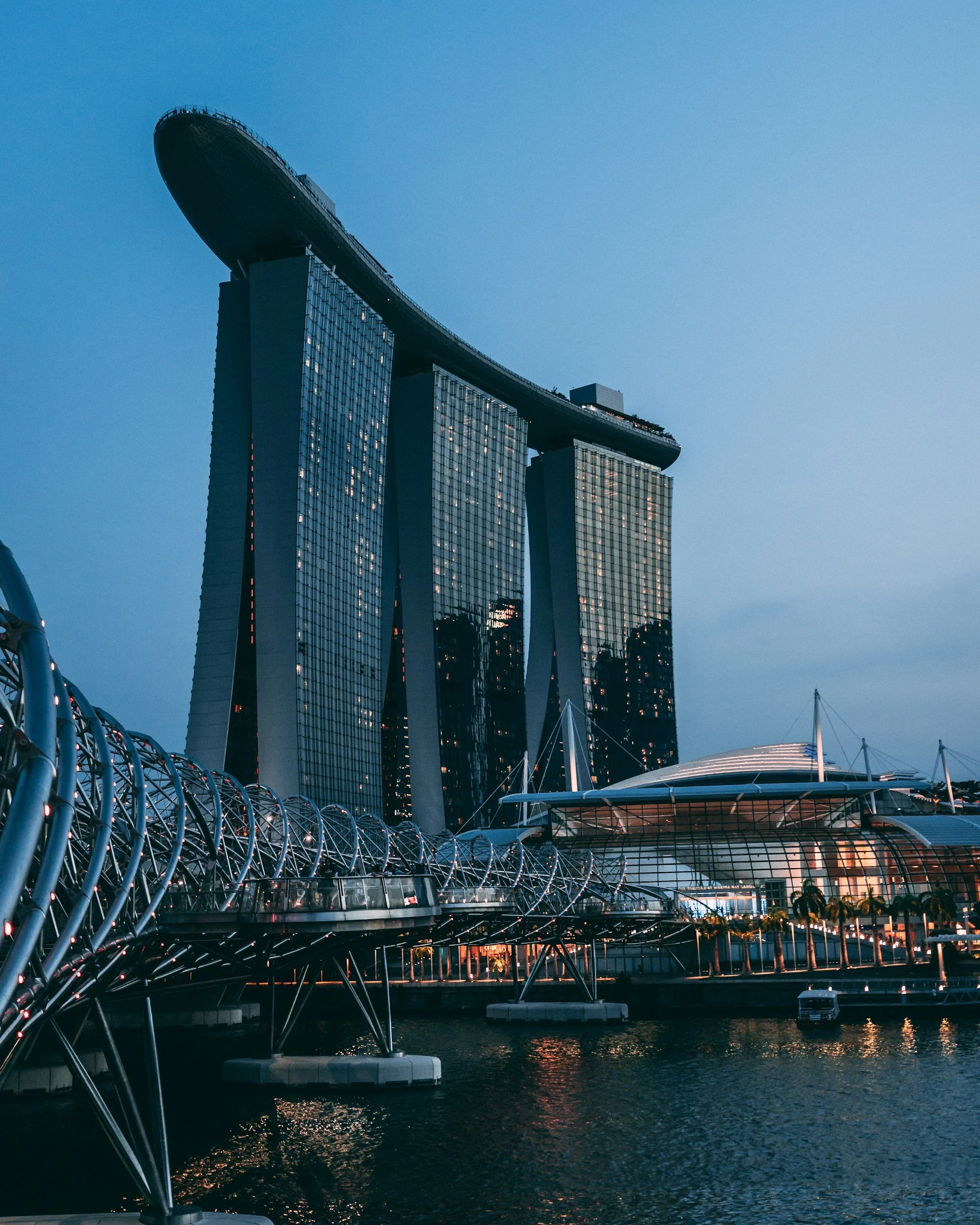 Night view of the Marina Bay Sands hotel in Singapore with its three towers topped by a boat-shaped structure, a curved pedestrian bridge in the foreground, and the waterfront below.