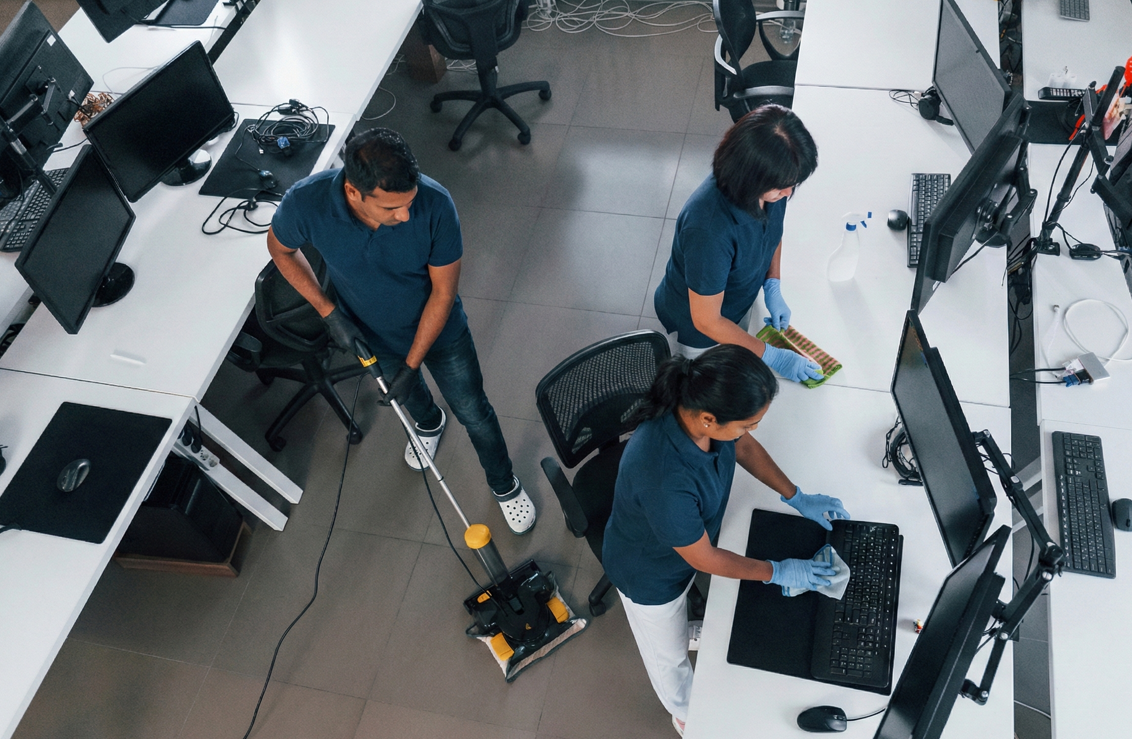 Three people cleaning computer desks and monitors in an office.