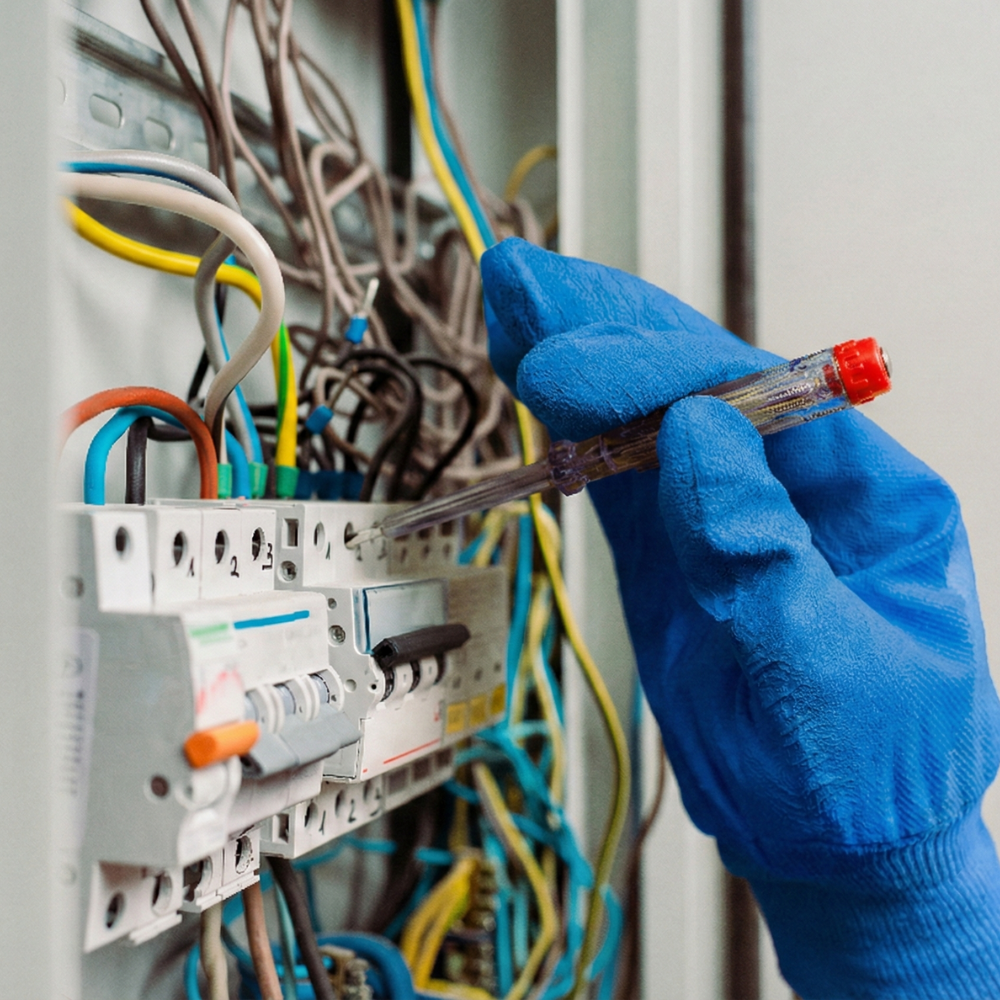A person wearing a blue glove using a screwdriver to work on an electrical panel with multiple wires and circuit breakers inside.