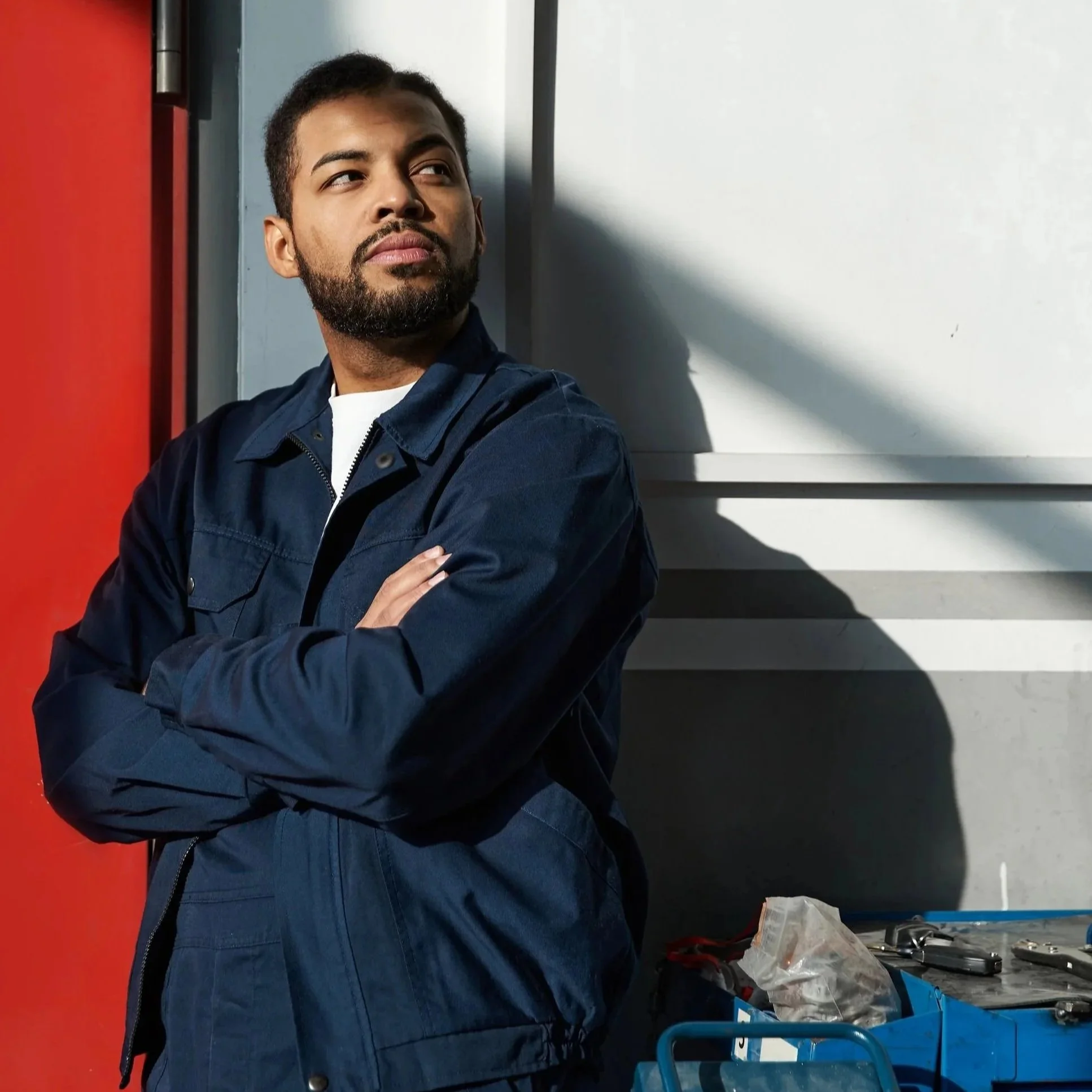 A man with a beard and mustache leaning against a wall, crossing his arms, looking to the side. He is wearing a dark blue jacket and standing next to a work cart filled with tools.