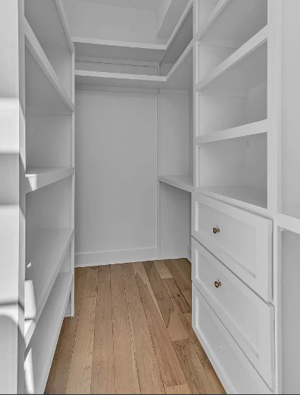 Empty white walk-in closet with built-in shelves and drawers, and light wood flooring.