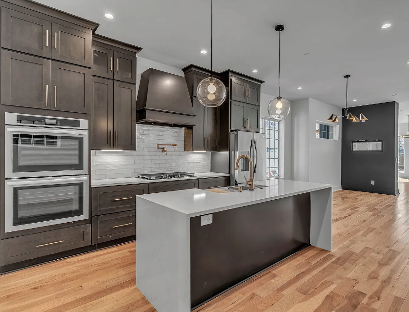 Modern kitchen with dark wood cabinets, white subway tile backsplash, and a white kitchen island with a gold faucet. Pendant lights hang above the island, and there is a stainless steel double oven and refrigerator. The flooring is light hardwood, an