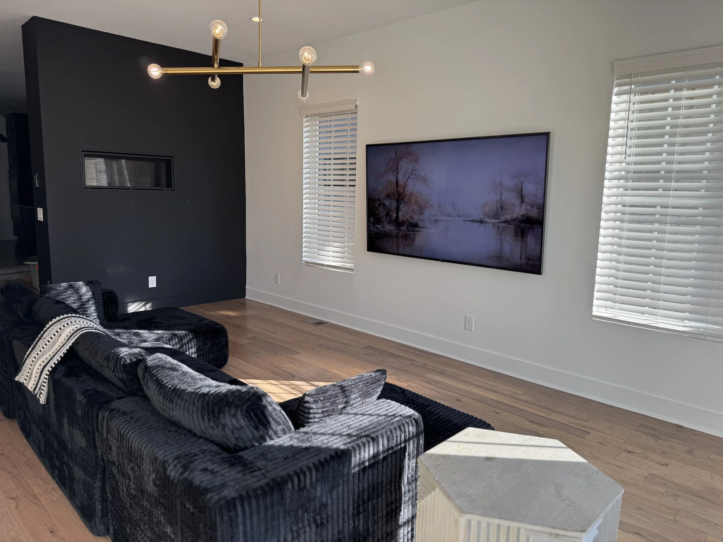 Living room with black velvet sofa, wooden floor, wall-mounted TV showing a landscape, and windows with white blinds.