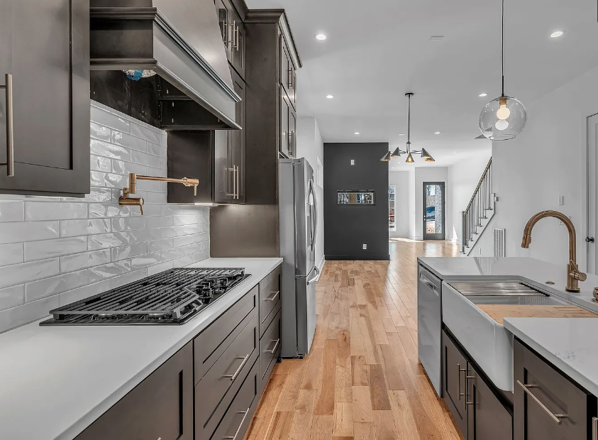 Modern kitchen with dark gray cabinetry, white subway tile backsplash, stainless steel appliances, wooden floors, and a kitchen island with a sink and brass faucet. Open layout leads to living area and staircase.
