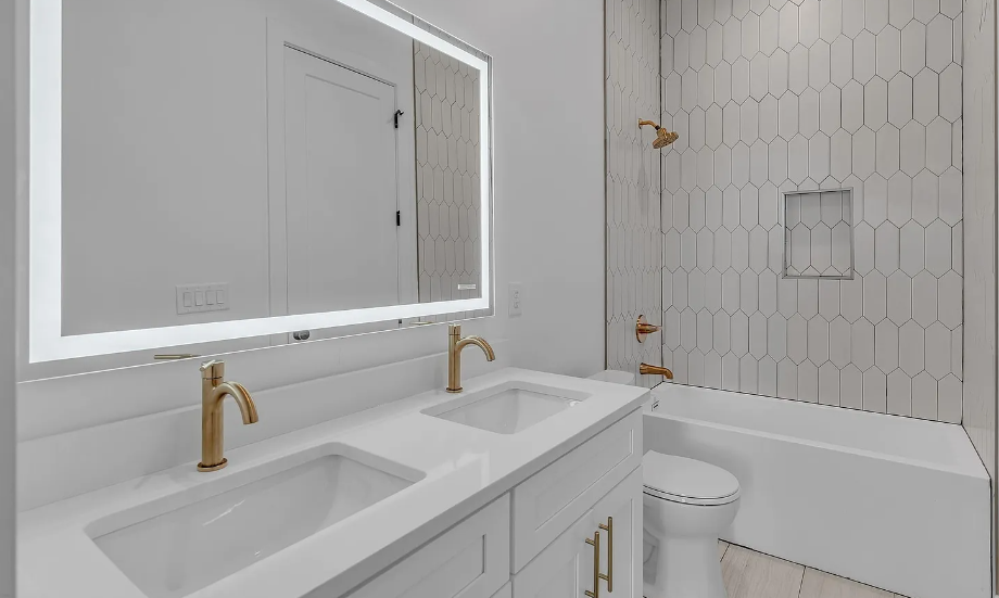 Modern bathroom with dual sinks, large illuminated mirror, and a bathtub with a showerhead. The walls are tiled in white with a honeycomb pattern, and the fixtures are gold.