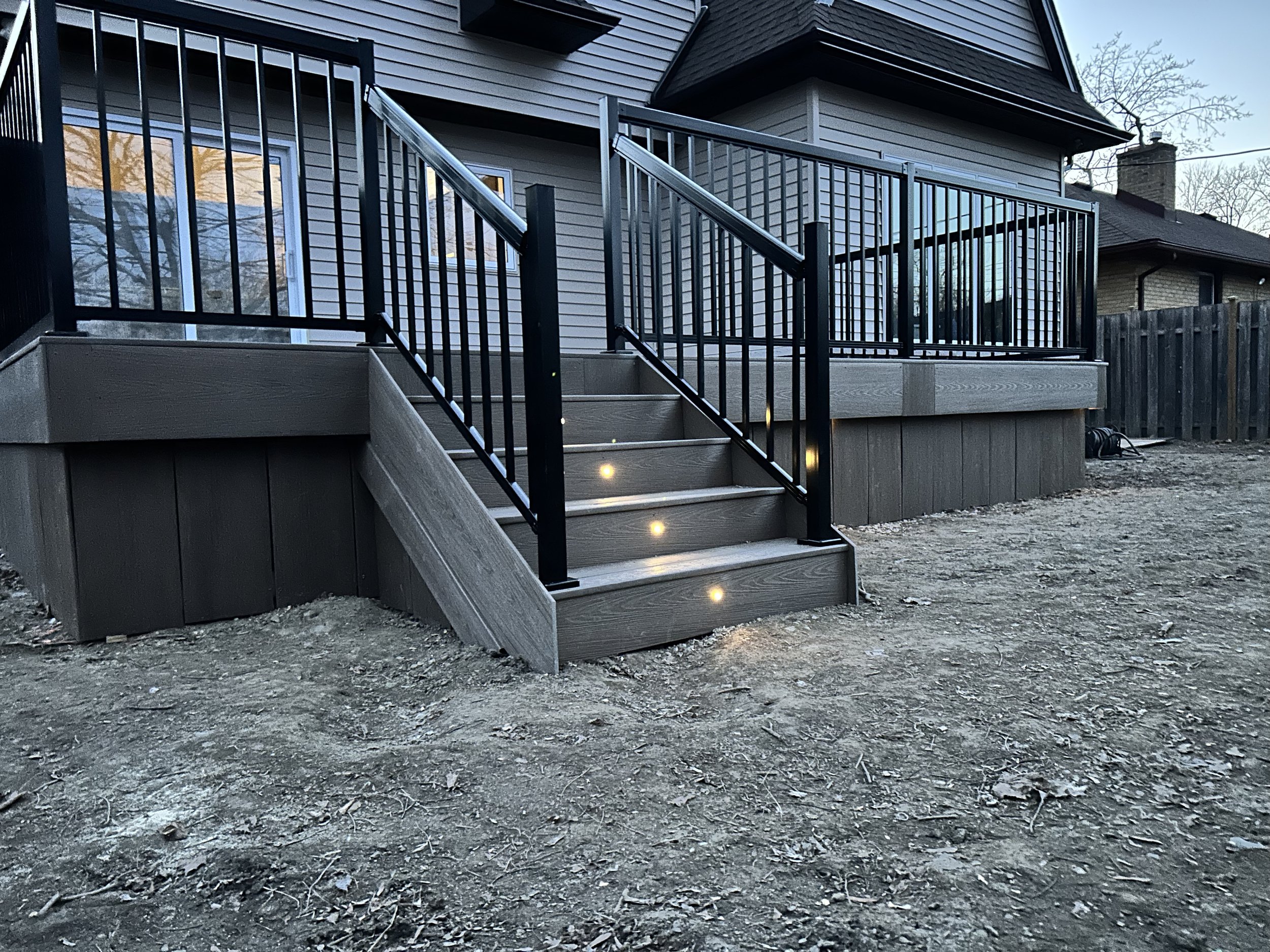Rear view of a house deck with black metal railing and built-in lights on the stairs, with a dirt yard in the foreground and neighboring houses in the background.
