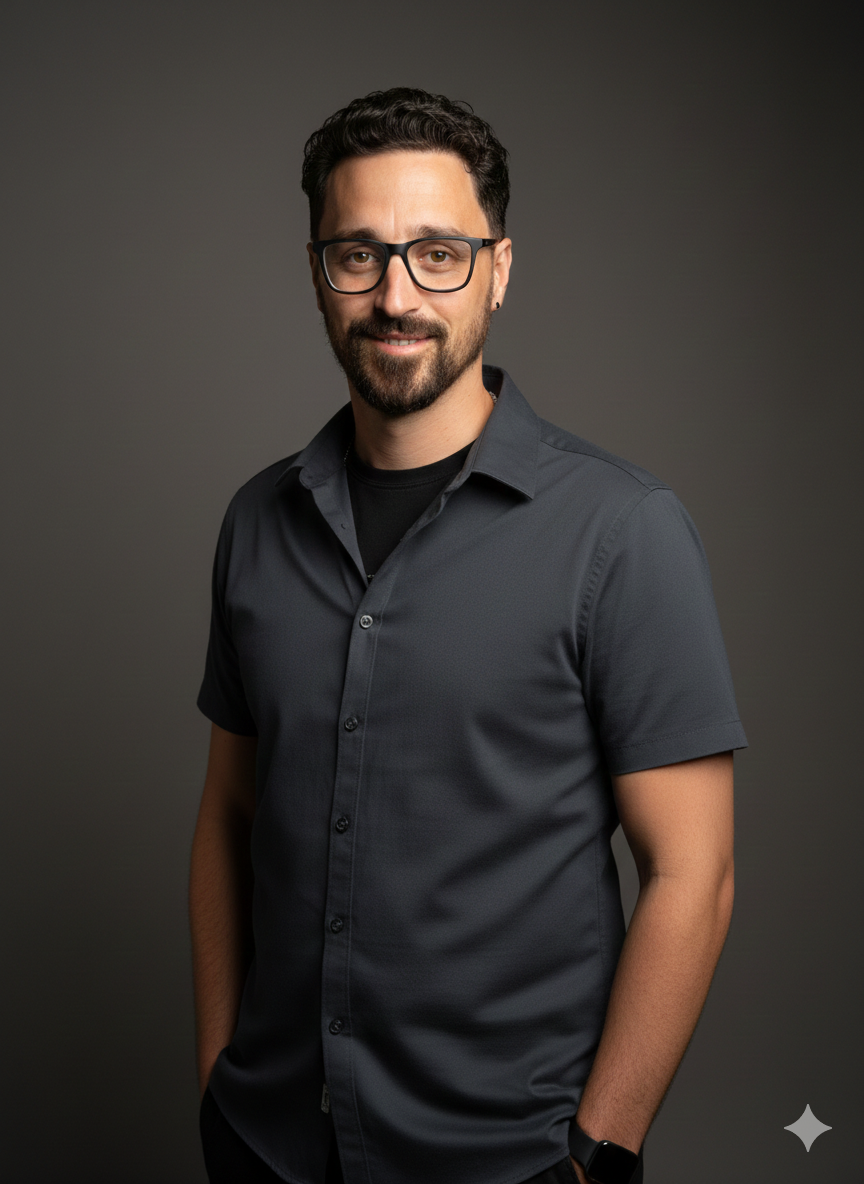 Portrait of a man with dark hair, beard, black glasses, wearing a dark gray button-up shirt, smiling against a dark gray background.