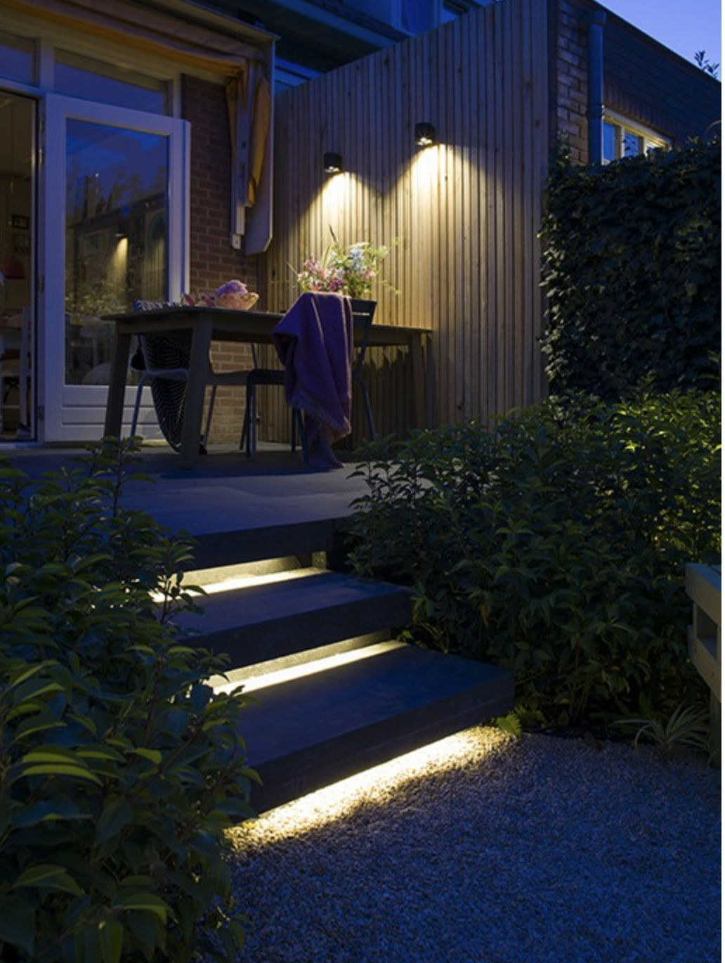 A backyard patio at night with three steps leading up to a wooden deck. The deck has a table with chairs, a vase of flowers, and some items on it. Two wall-mounted lights illuminate the area, and the surrounding bushes are visible in the dark.