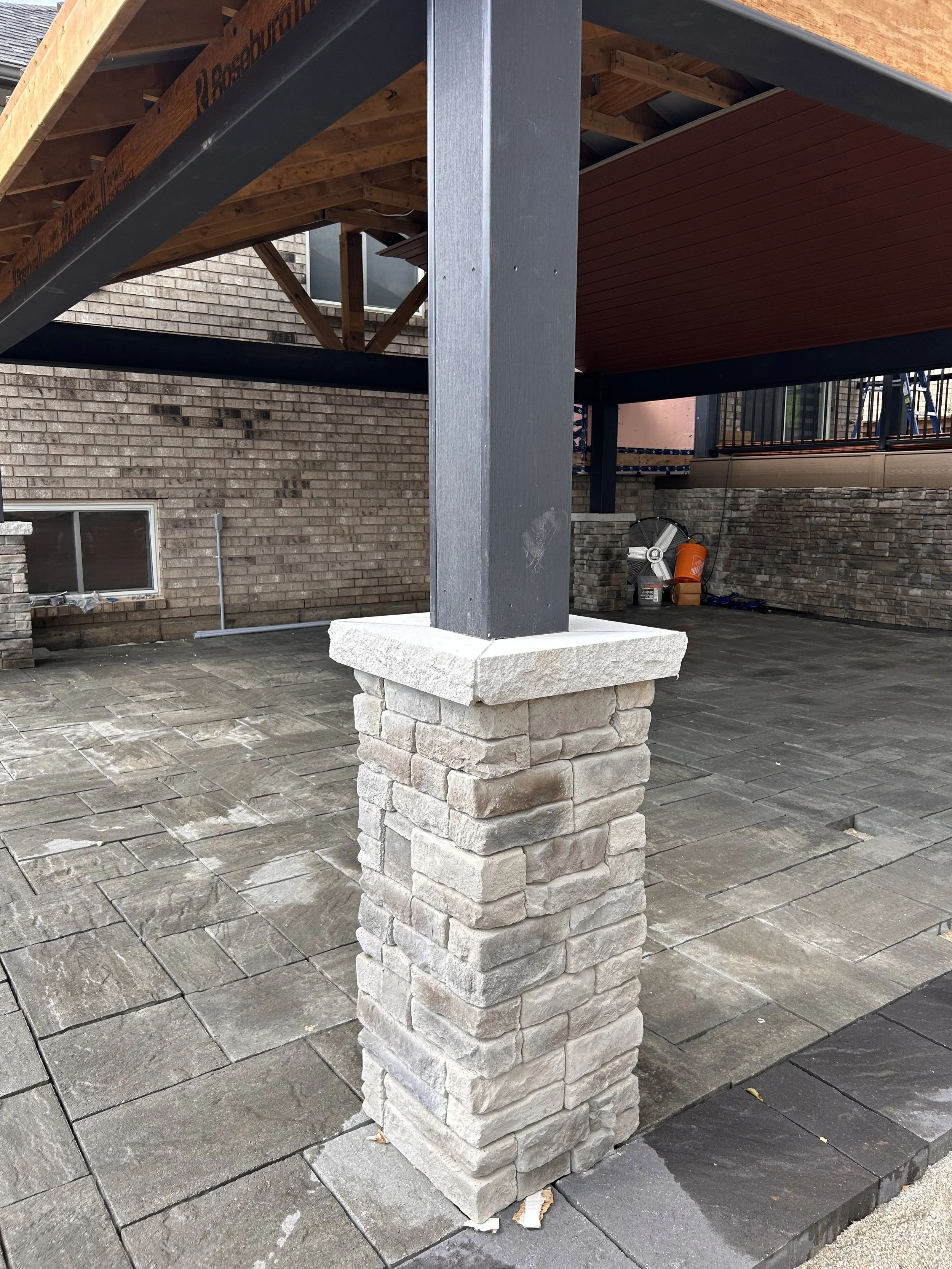 Close-up view of a stone pillar supporting a gazebo, with a brick building and outdoor tools in the background.