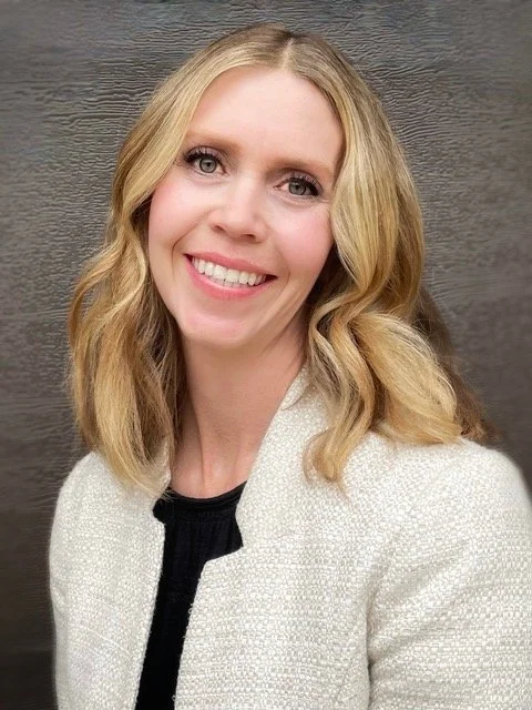 A smiling woman with blonde wavy hair, wearing a light-colored blazer over a black top, against a dark wooden background.