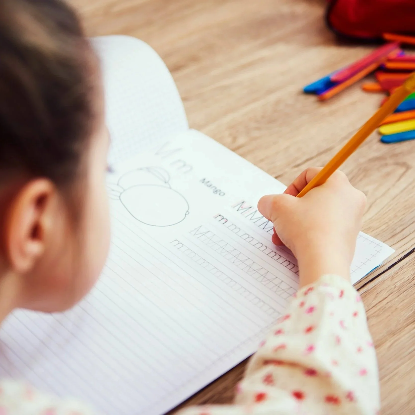 A child practicing writing the letter M in a workbook, with colored markers on a wooden table.