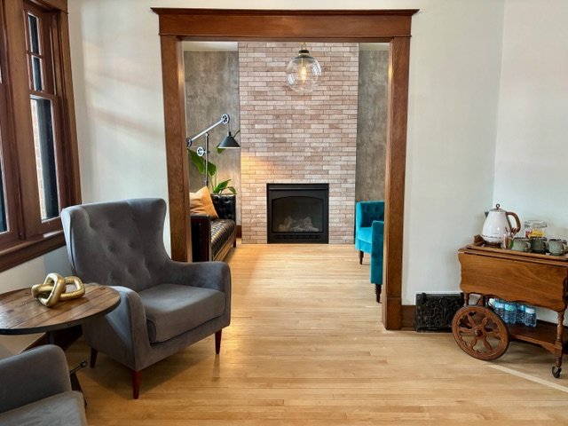 Living room with wood flooring, a gray armchair, a wooden side table with gold decorative objects, a stone fireplace, and a brick accent wall. There is a wooden doorway framing the view into the adjoining room.