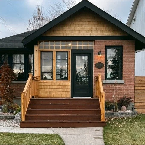 Front view of a house with a small porch, black door, and wooden stairs, combining brick and wood siding.
