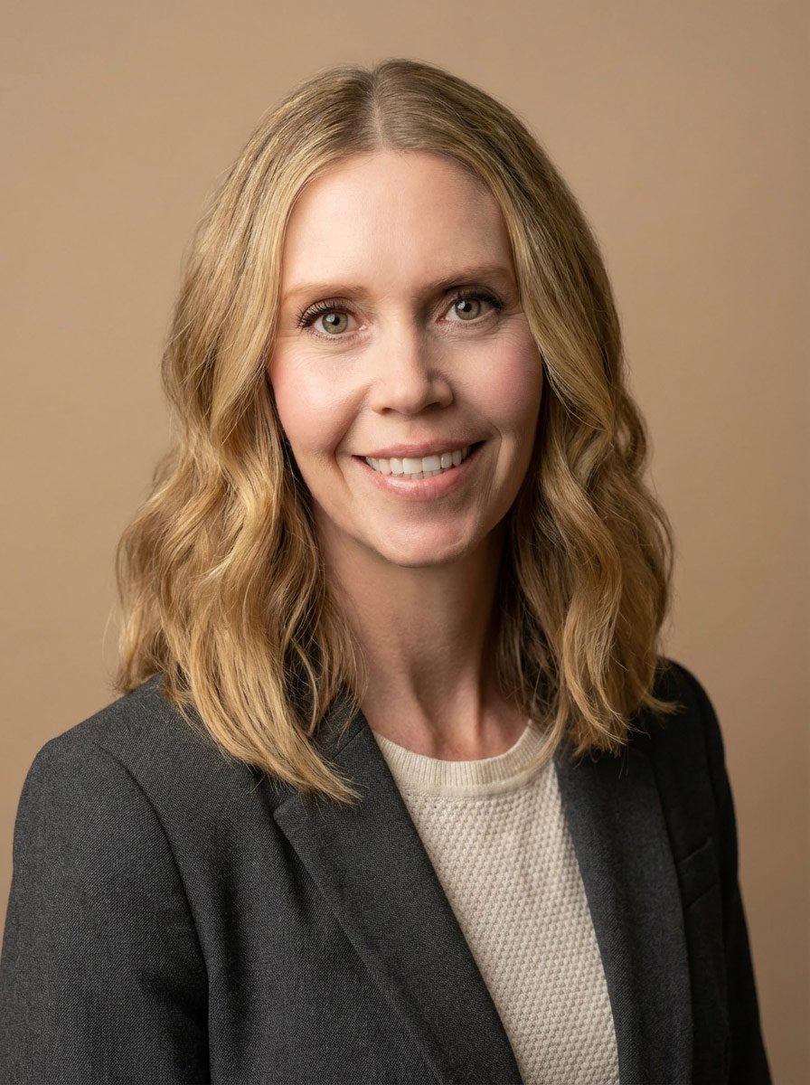 A woman with shoulder-length blonde hair, wearing a black blazer and a beige sweater, smiling against a beige background.