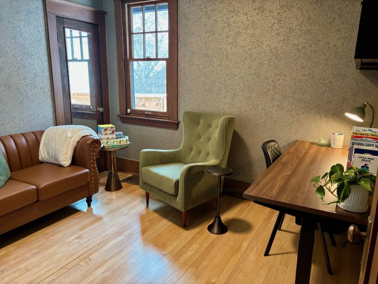 A cozy living room corner with a brown leather sofa, a light green high-back armchair, a wooden side table with colorful puzzles, and a wooden desk with a lamp, a plant, and some children's books. There is a window and a door with wooden frames, and hardwood flooring.