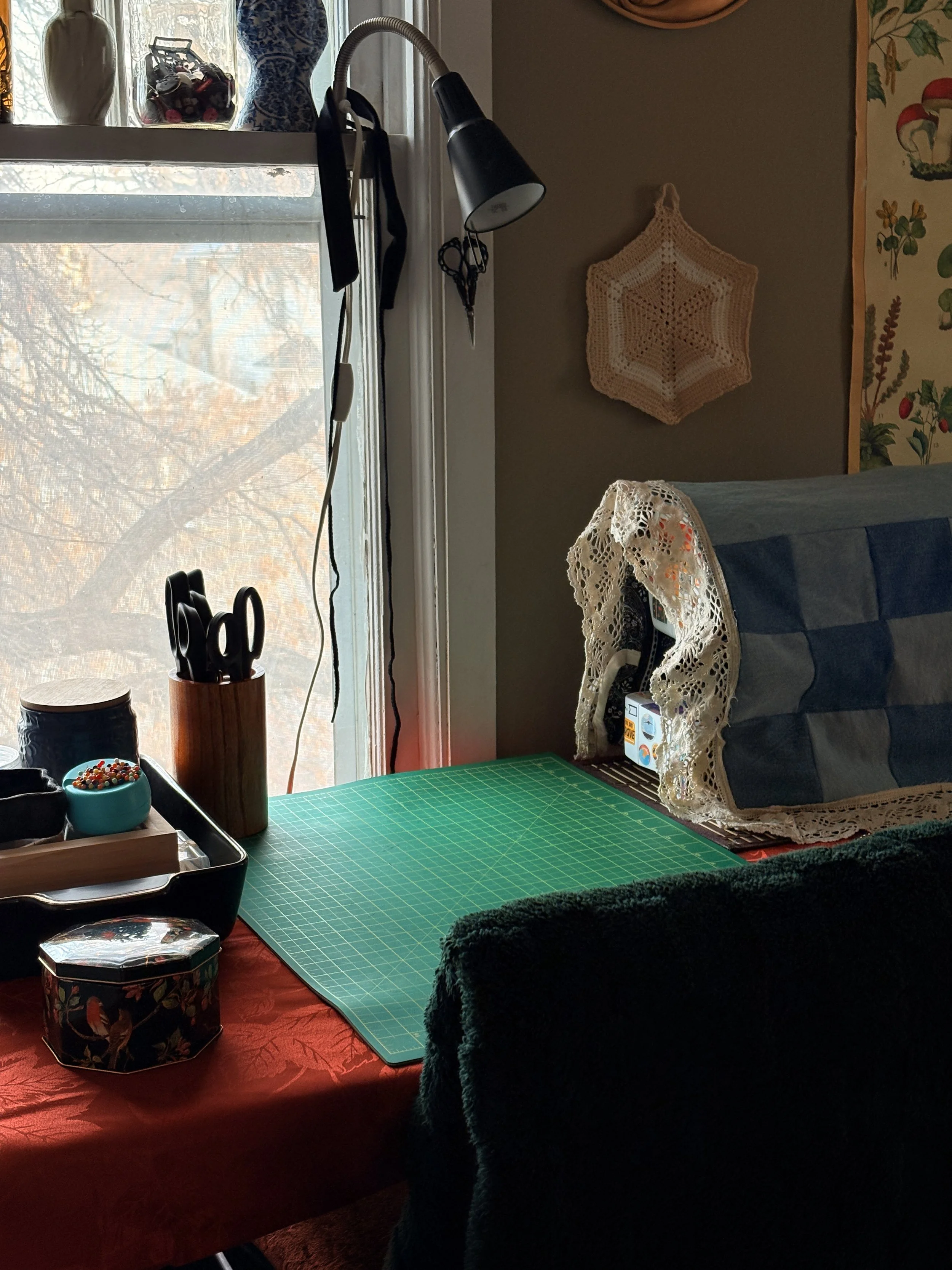 A craft workspace with a green cutting mat on a red table. Various tools and containers are on the table, with a window showing trees outside. A paper craft project with blue and white polka dots is on a cloth-covered object, and a crocheted wall decoration is visible on the wall.
