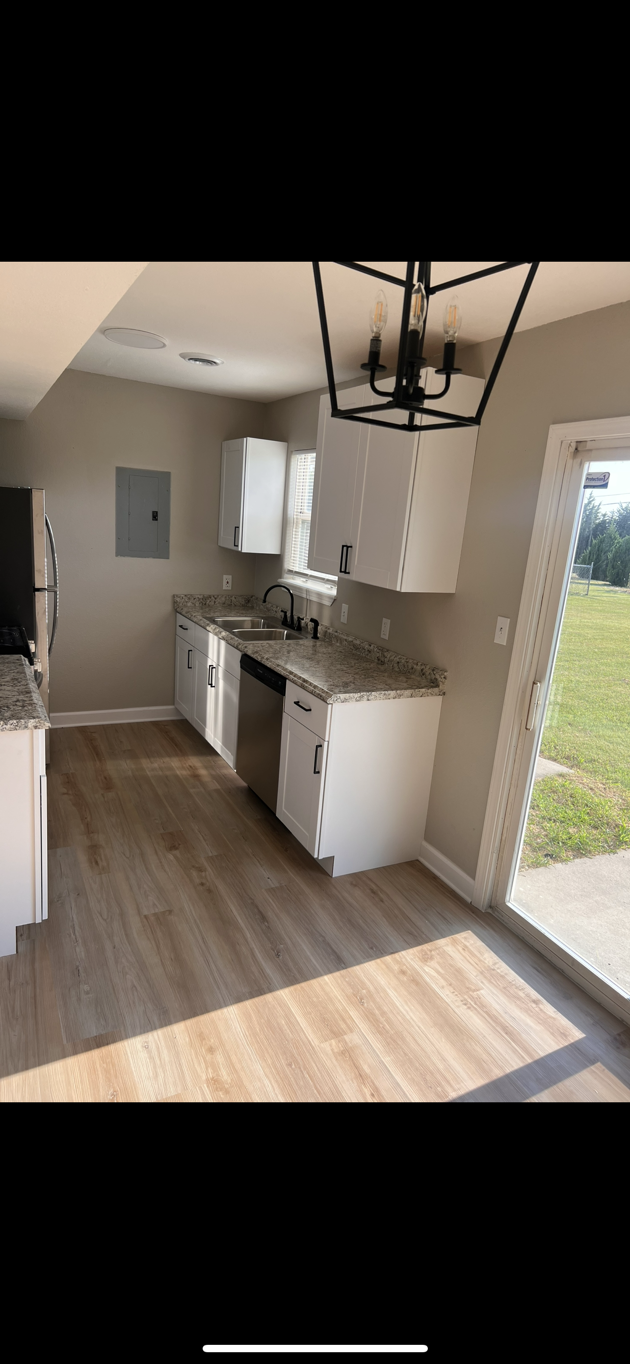 Kitchen with white cabinets, granite countertops, stainless steel dishwasher, black faucet, window, sliding glass door opening to outdoor yard, light wood flooring, black geometric chandelier.