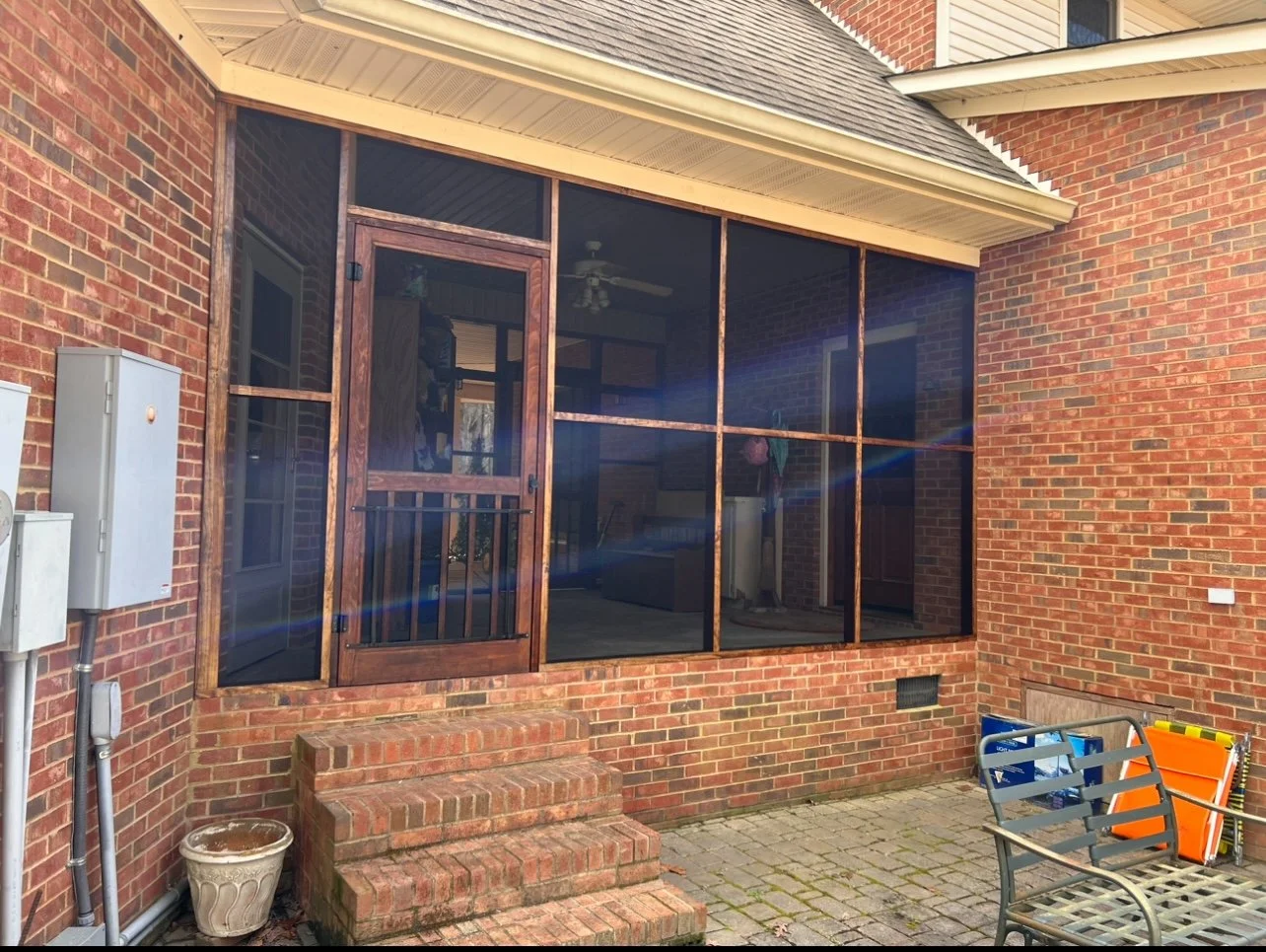 Back porch area with brick steps leading up to a screened-in porch with a wooden door. The porch has a brick foundation and a roof extension. There's outdoor seating and storage boxes, along with utility meters and a vent on the wall.