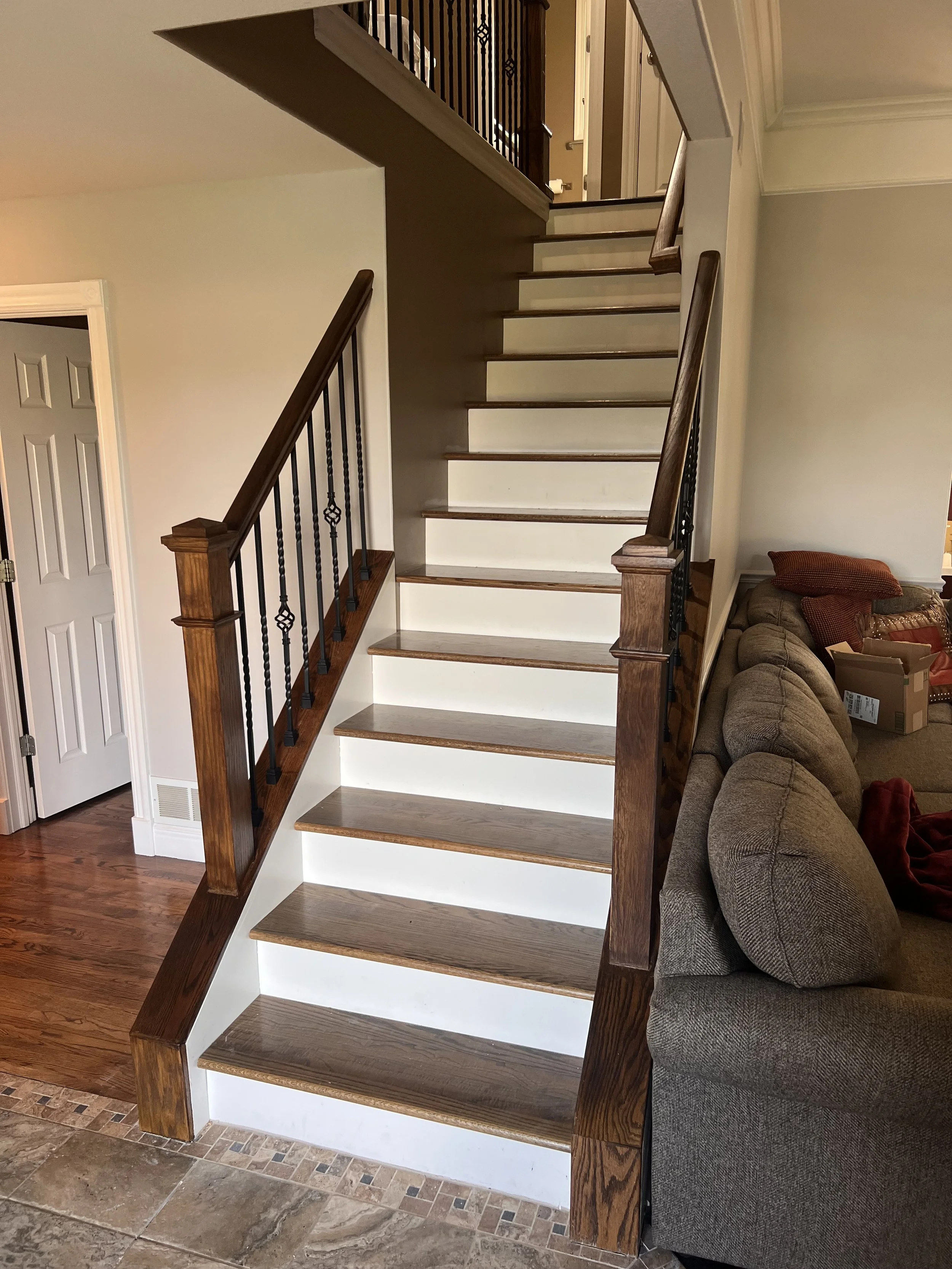 Indoor staircase with wooden steps, dark wood handrails, and black metal balusters, leading to an upper floor, with a living room on the right and a door on the left.