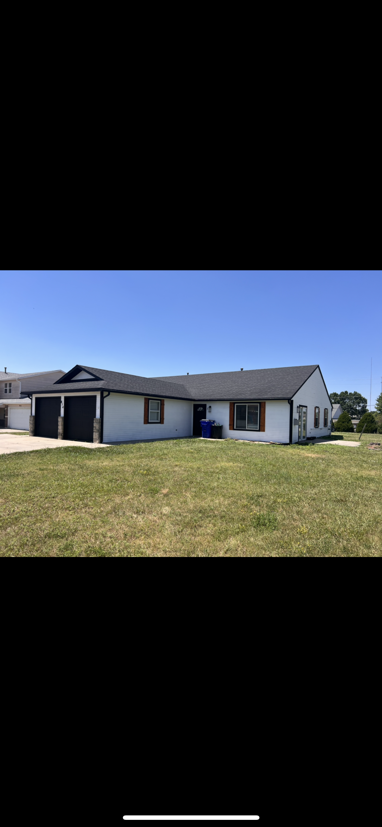Single-story house with white siding, dark roof, two garage doors, wooden shutters, front door, and a grassy yard under a clear blue sky.