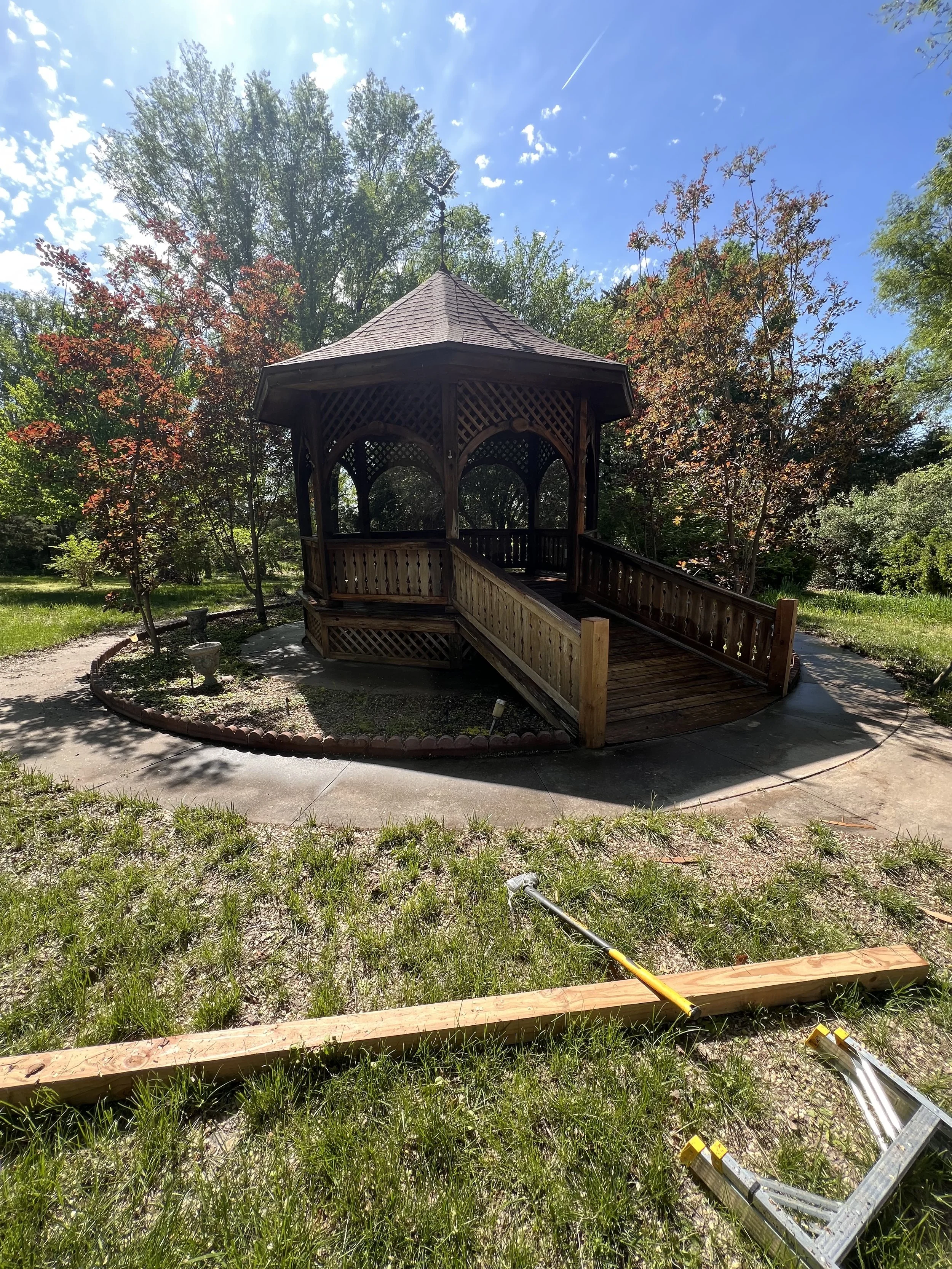 A gazebo in a park surrounded by trees on a sunny day, with tools and wood pieces on the grass nearby, possibly for repair or construction.