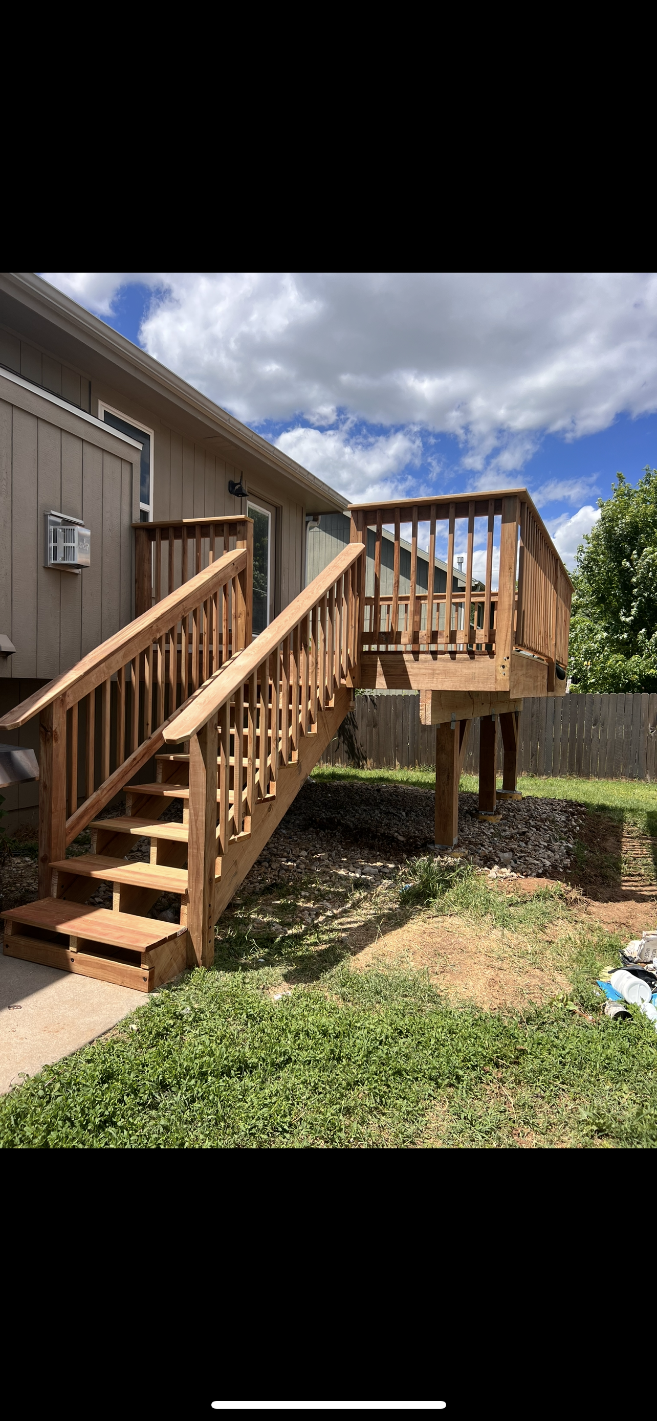 Newly built wooden deck with stairs attached to a house, set in a backyard with green grass, under a blue sky with clouds.