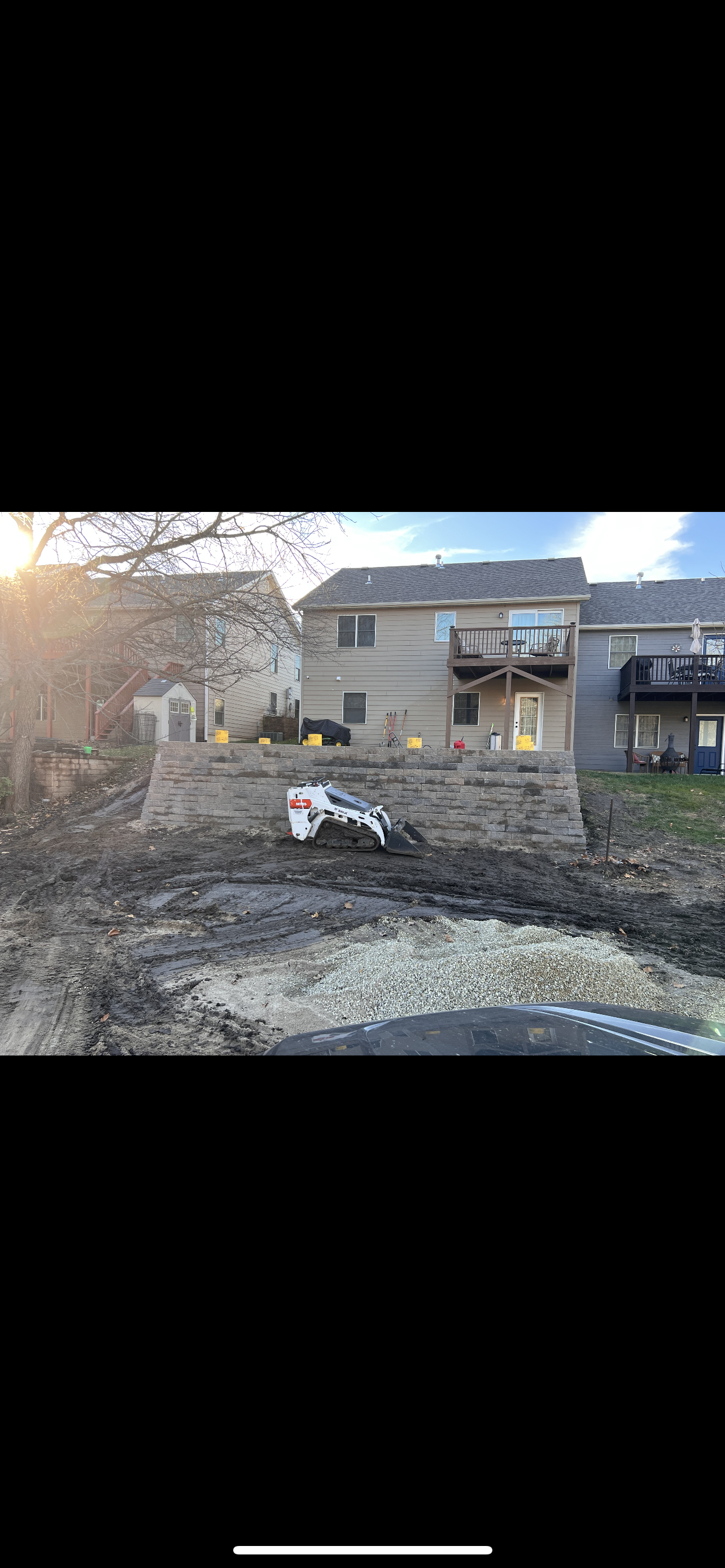 A construction site with a small white bulldozer working on leveling ground next to a stone retaining wall in a residential area, with houses and decks in the background.