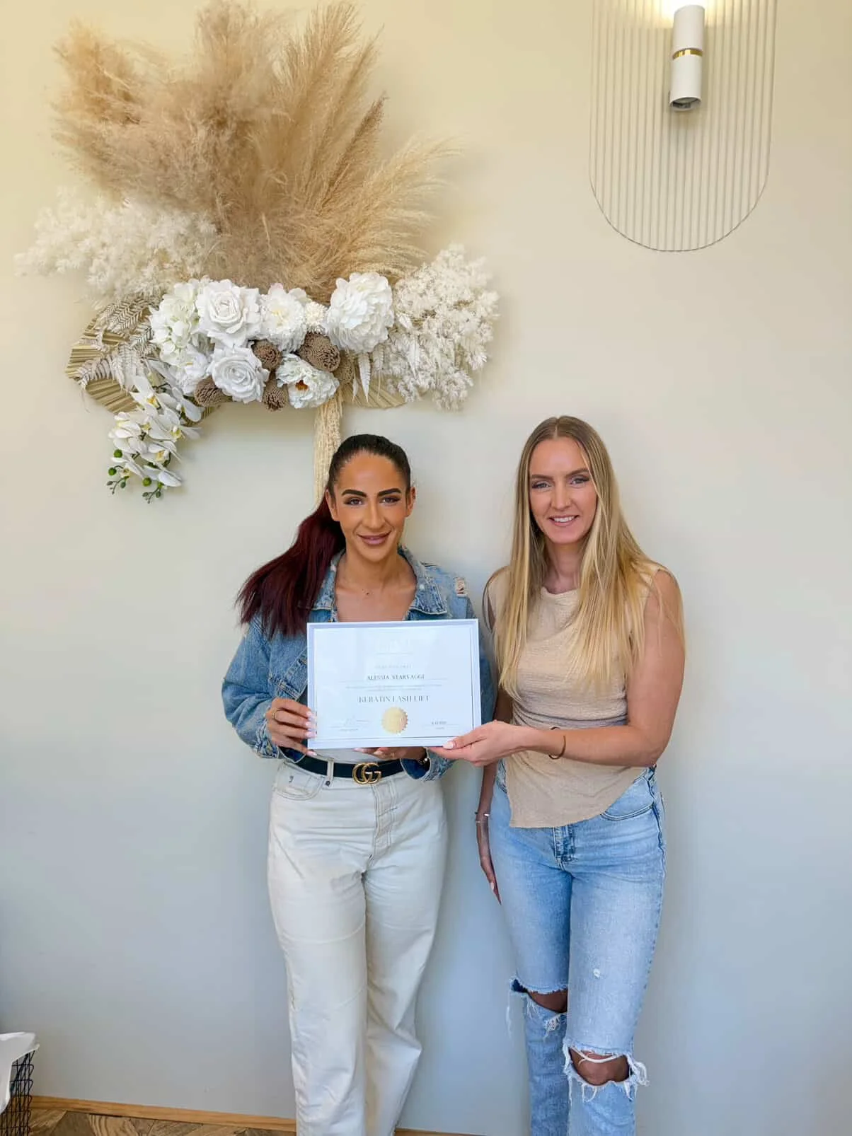 Two women standing side by side, one holding a certificate, in front of a wall decorated with a large floral and pampas grass wall art piece.