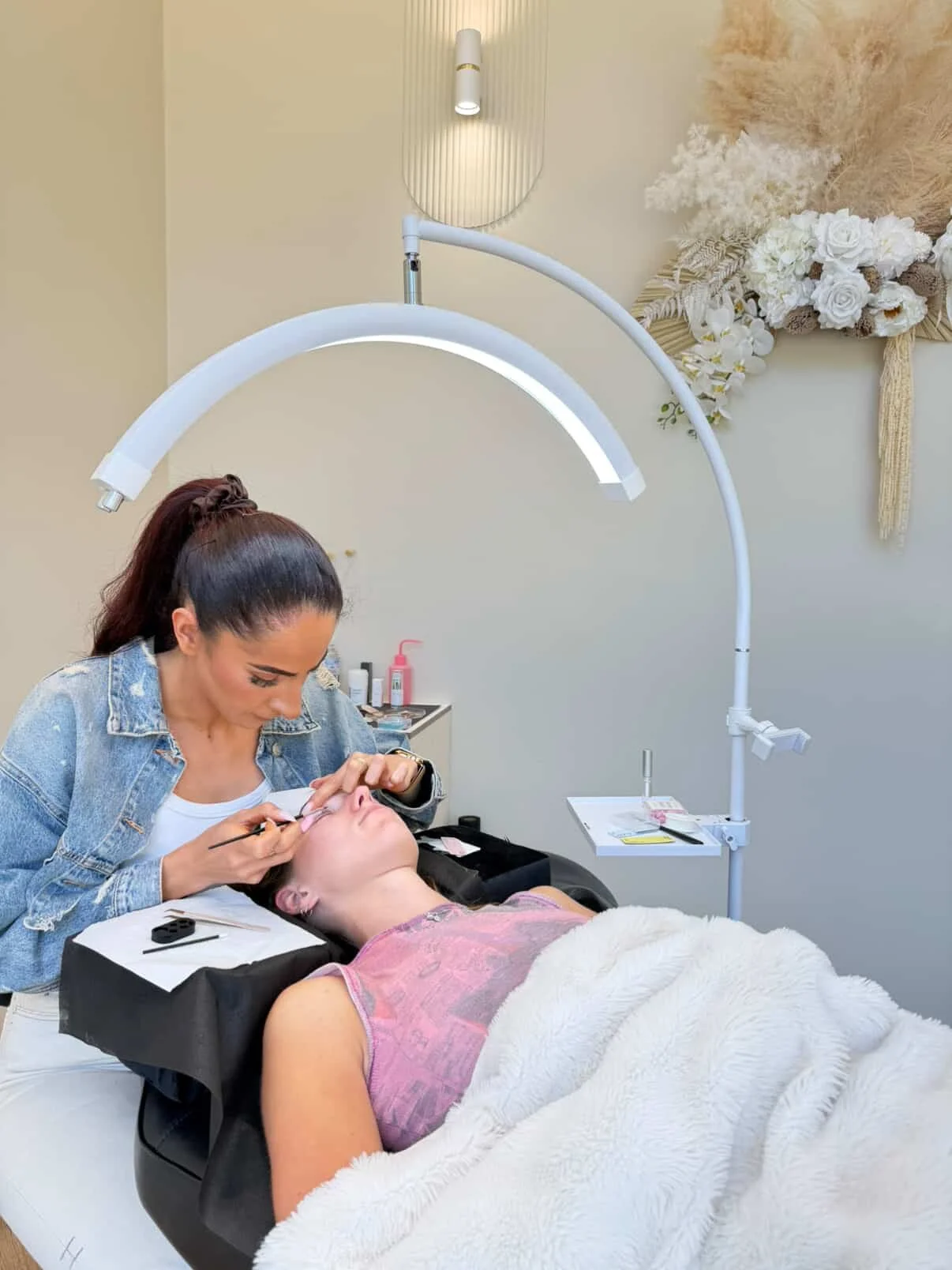 A woman receiving a cosmetic procedure on her face from a professional in a spa-like setting, with a large, curved overhead light and decorative flower wall in the background.