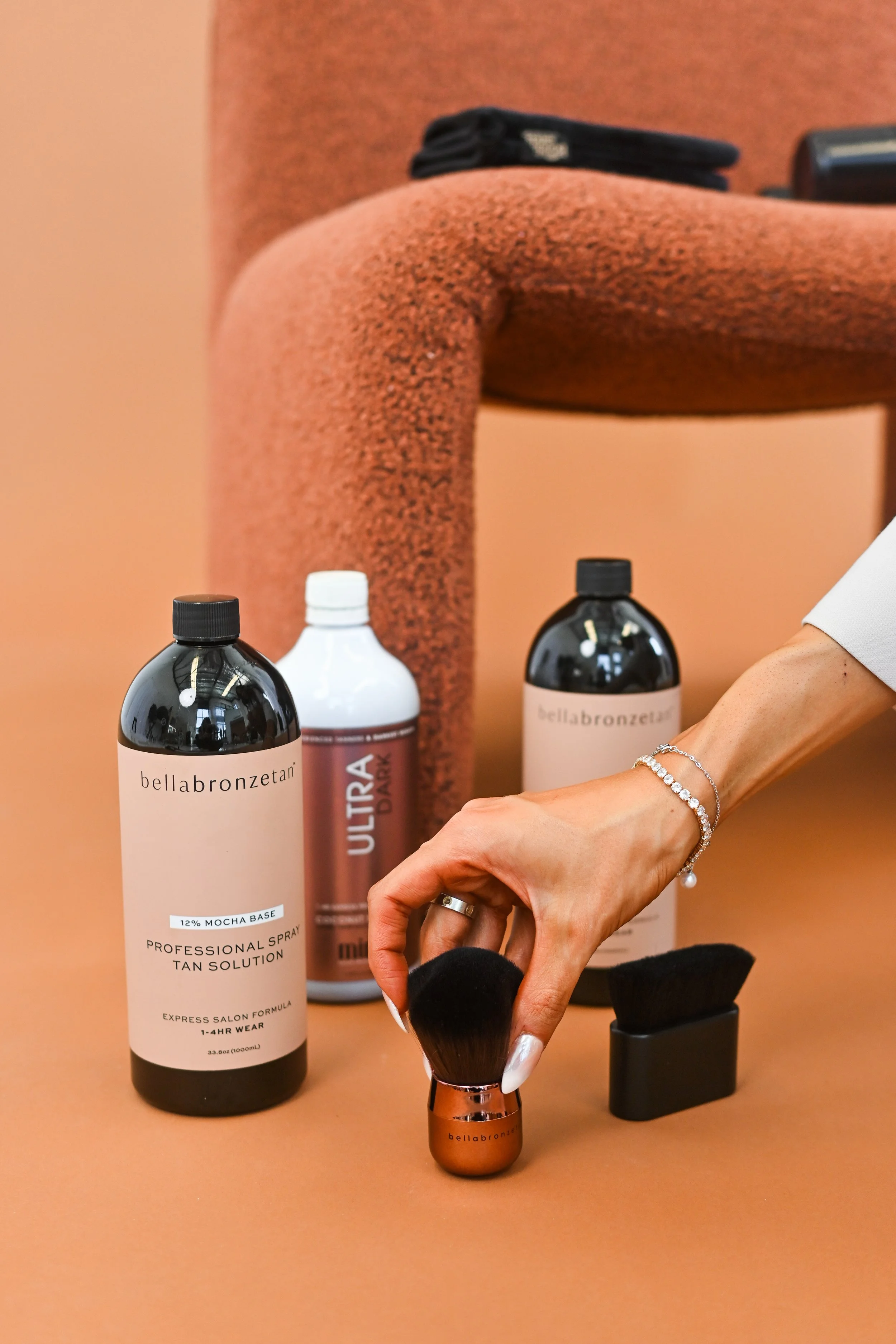 A person's hand holding a makeup brush near bottles of tanning products on a tan-colored table, with a textured tan armrest in the background.