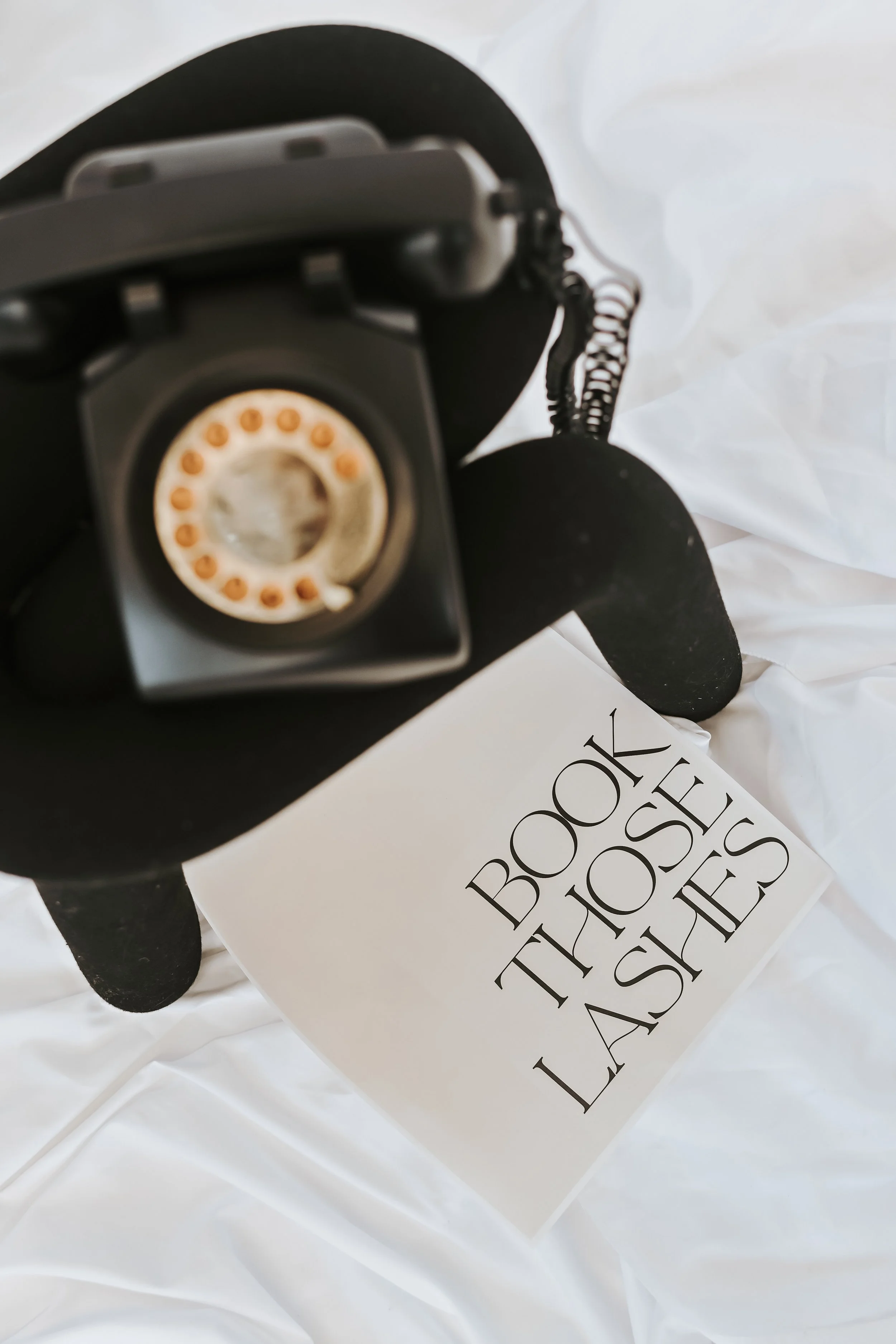 A vintage film camera mounted on a tripod, with a book titled "BOOK THE MOST LASHES" beside it on white fabric.