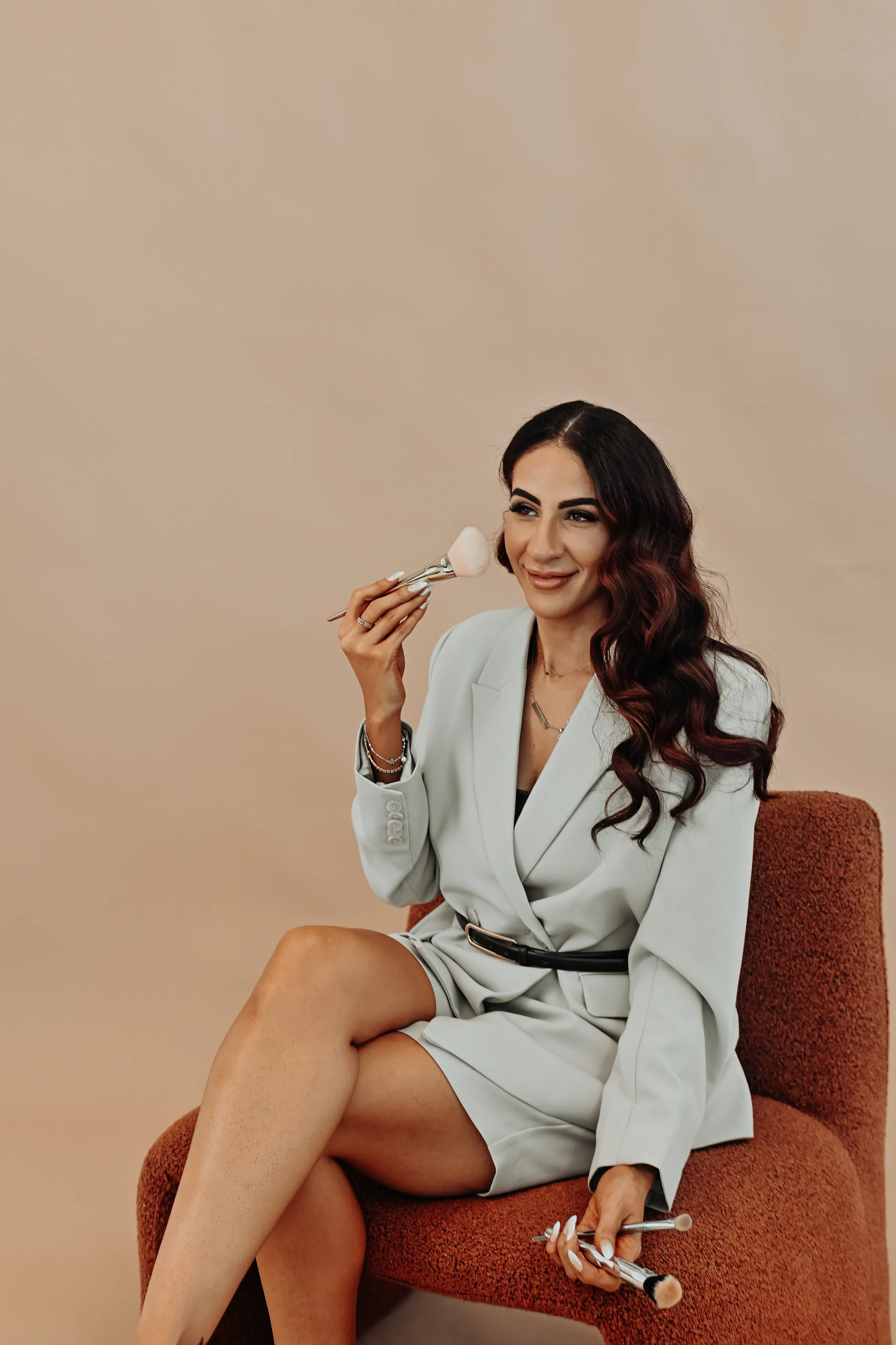 A woman sitting on a rust-colored chair, holding makeup brushes, smiling, with long dark hair and wearing a light-colored blazer dress.
