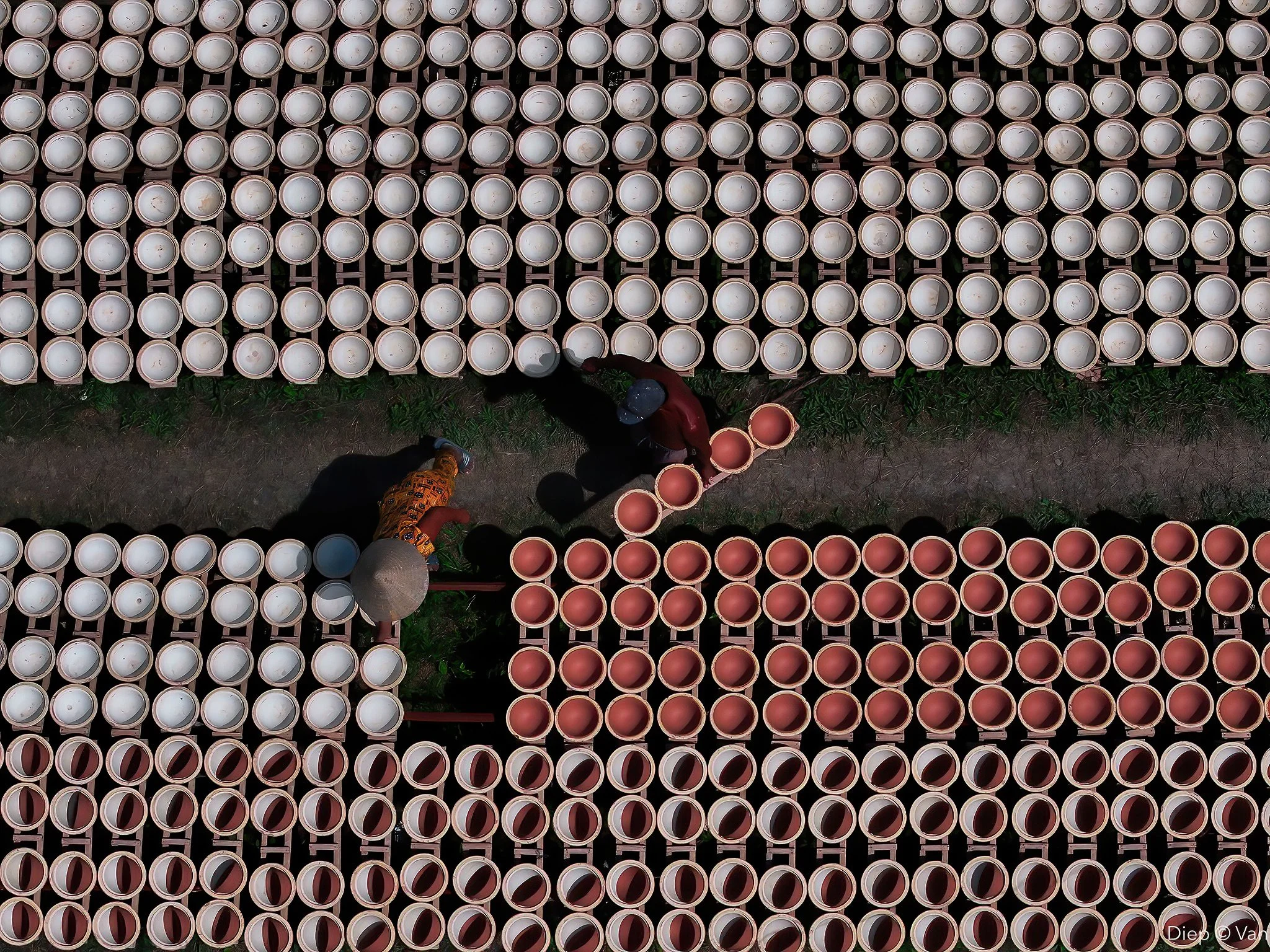 Two workers are stacking terracotta flower pots in a field, surrounded by white plastic buckets stacked on shelves.