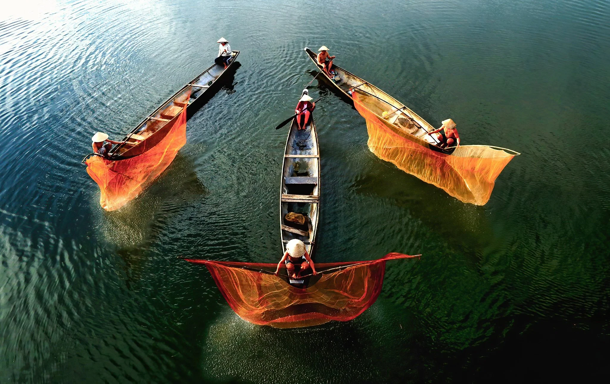 Five traditional boats with fishermen, some holding red fishing nets, on a calm greenish body of water, seen from above.