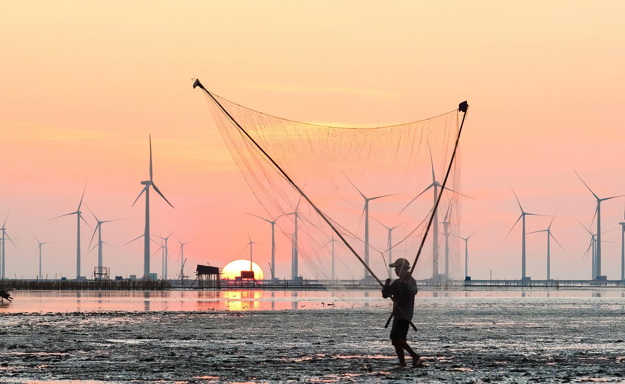A person walking on a sandy shore at sunset, carrying a large fishing net, with wind turbines in the background and the sun setting on the horizon.