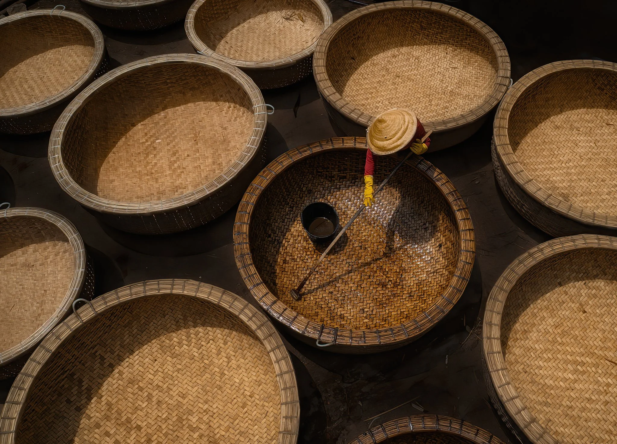 A person working in a workshop surrounded by woven baskets, using a tool to finish or clean the baskets.