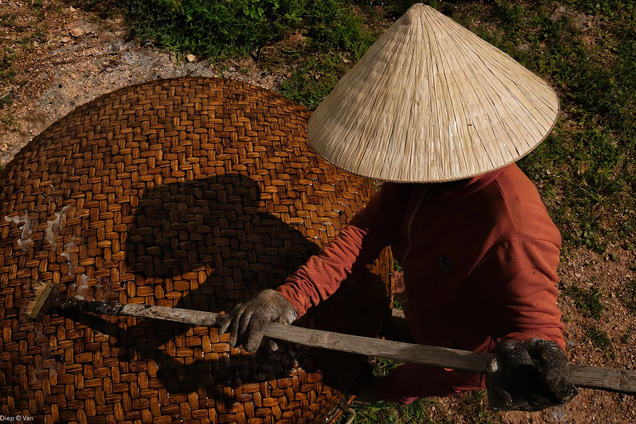 A person wears a traditional straw conical hat and work gloves, working with a large woven container made of natural materials, outdoors on a patch of dirt and grass.
