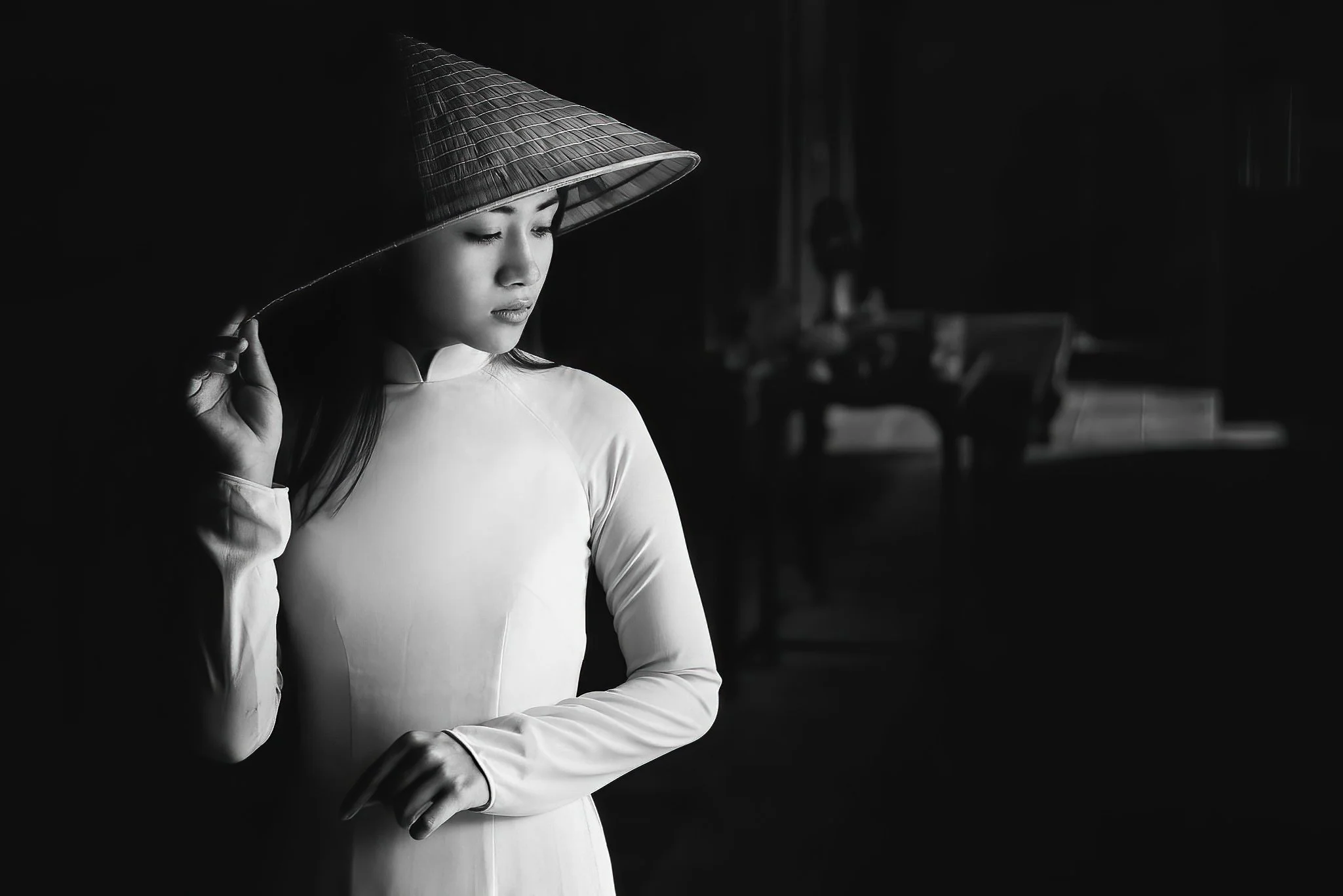 A woman in traditional Vietnamese attire wearing an áo dài and a non la, the conical hat, holding the brim of the hat with one hand and looking down, in a dimly lit room.