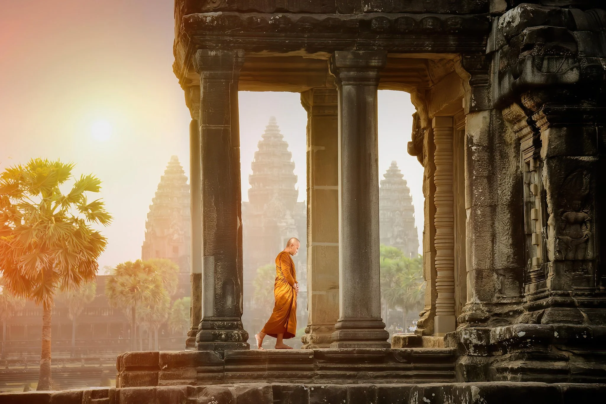 A Buddhist monk in orange robes walking through an ancient stone temple with tall columns, with the sun setting or rising in the background and palm trees nearby.