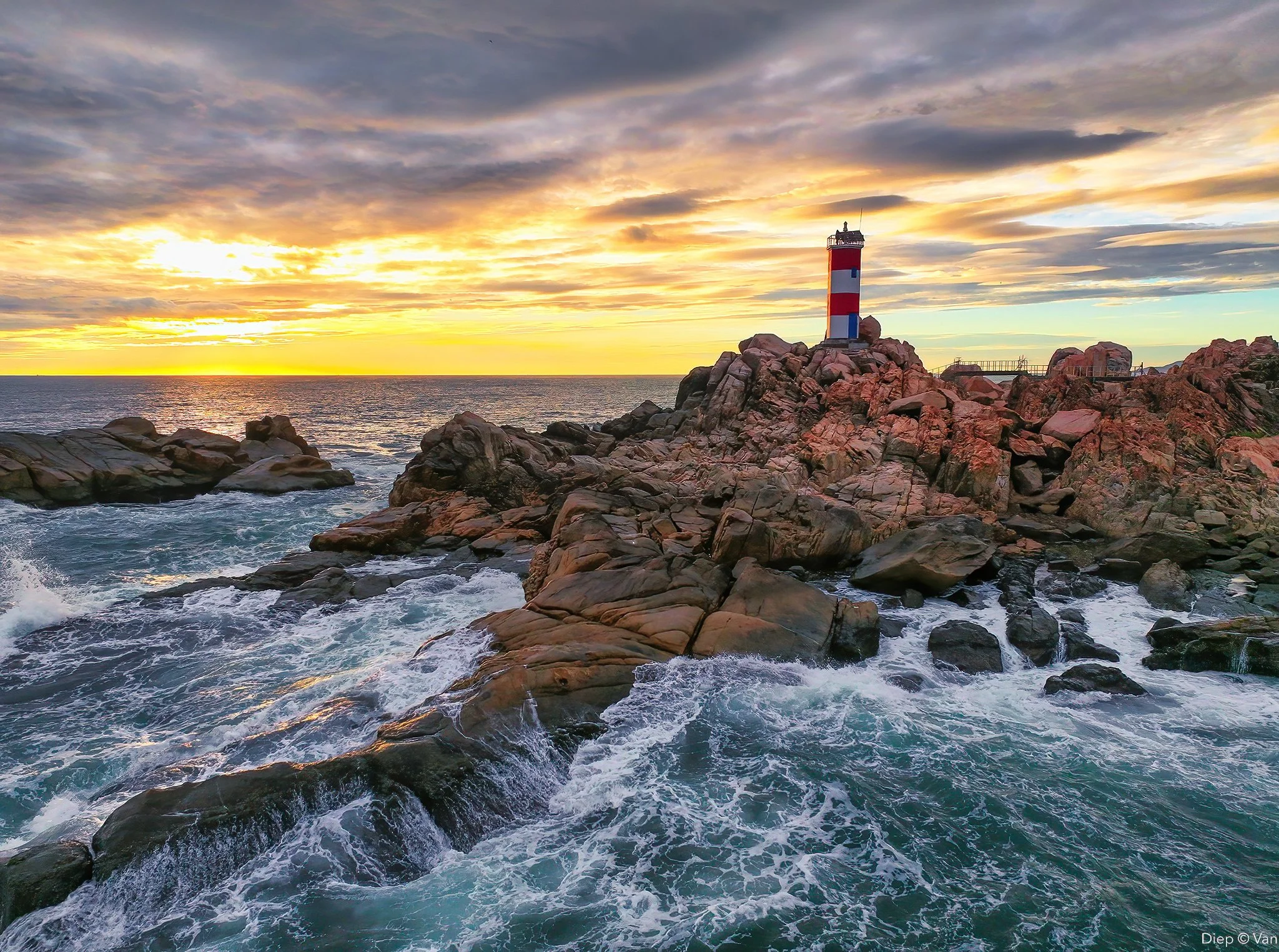 A lighthouse on rocky shores with waves crashing, overlooking the sunset over the ocean, and a cloudy sky.