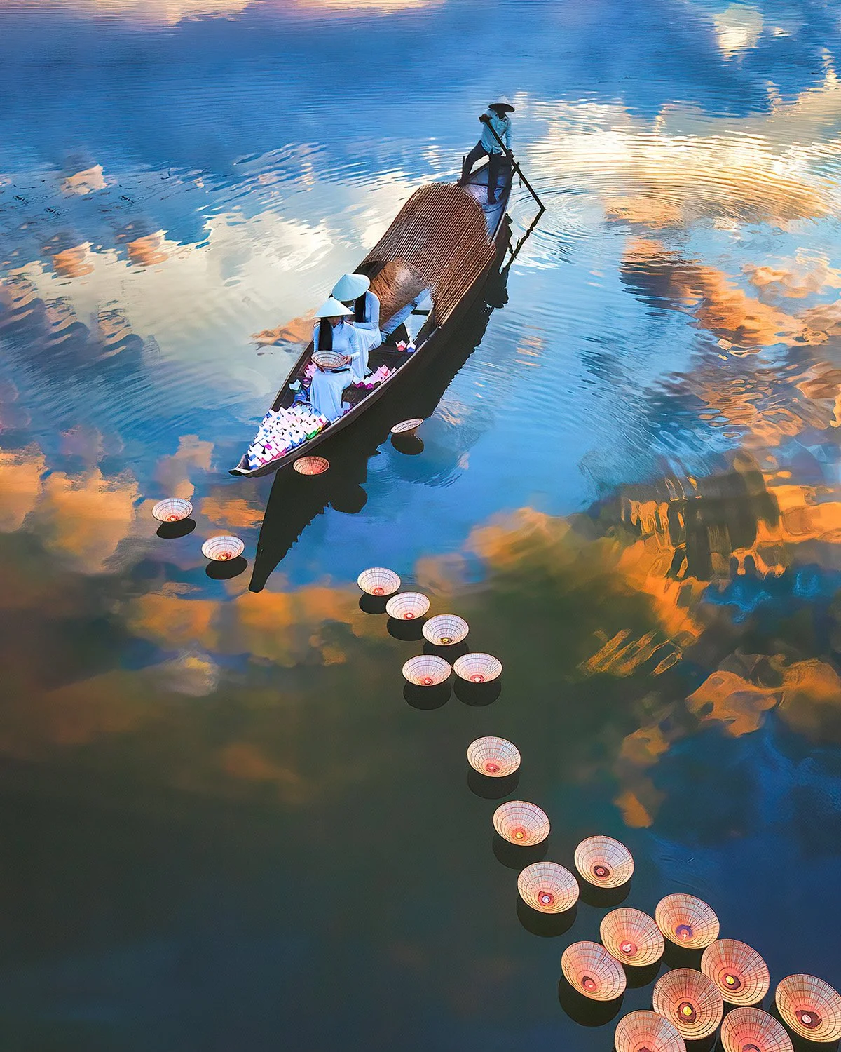 A boat carrying three women dressed in traditional Vietnamese Ao Dai and wearing conical hats, floating on a river with floating lanterns and reflected clouds and sky.