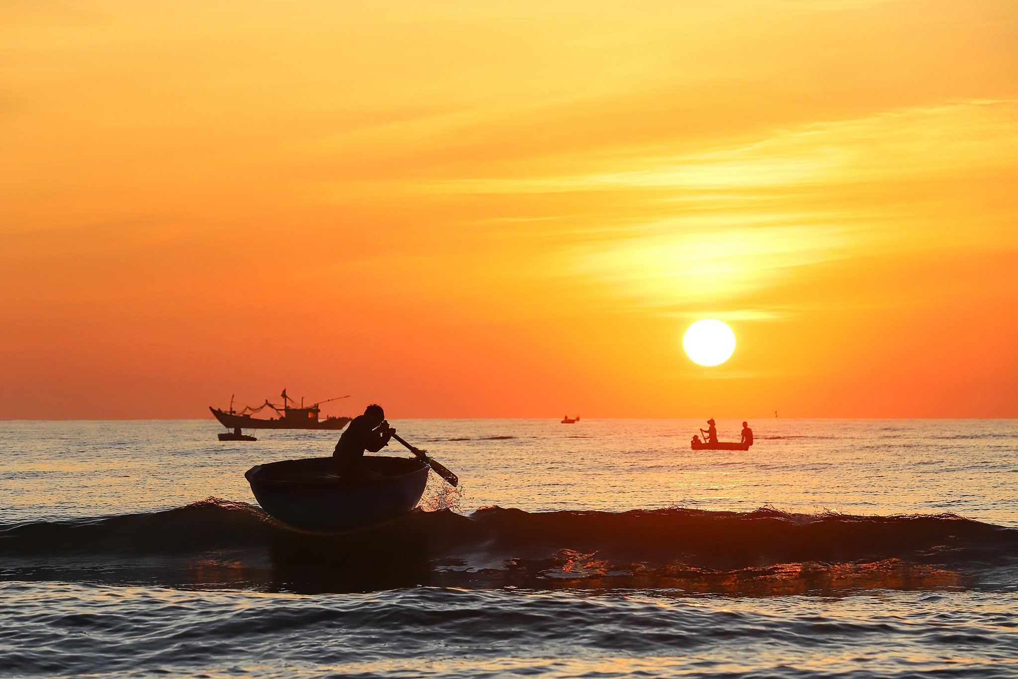 Silhouette of a person paddling a small boat on the ocean during a sunset, with several boats visible in the distance and a vibrant orange sky.