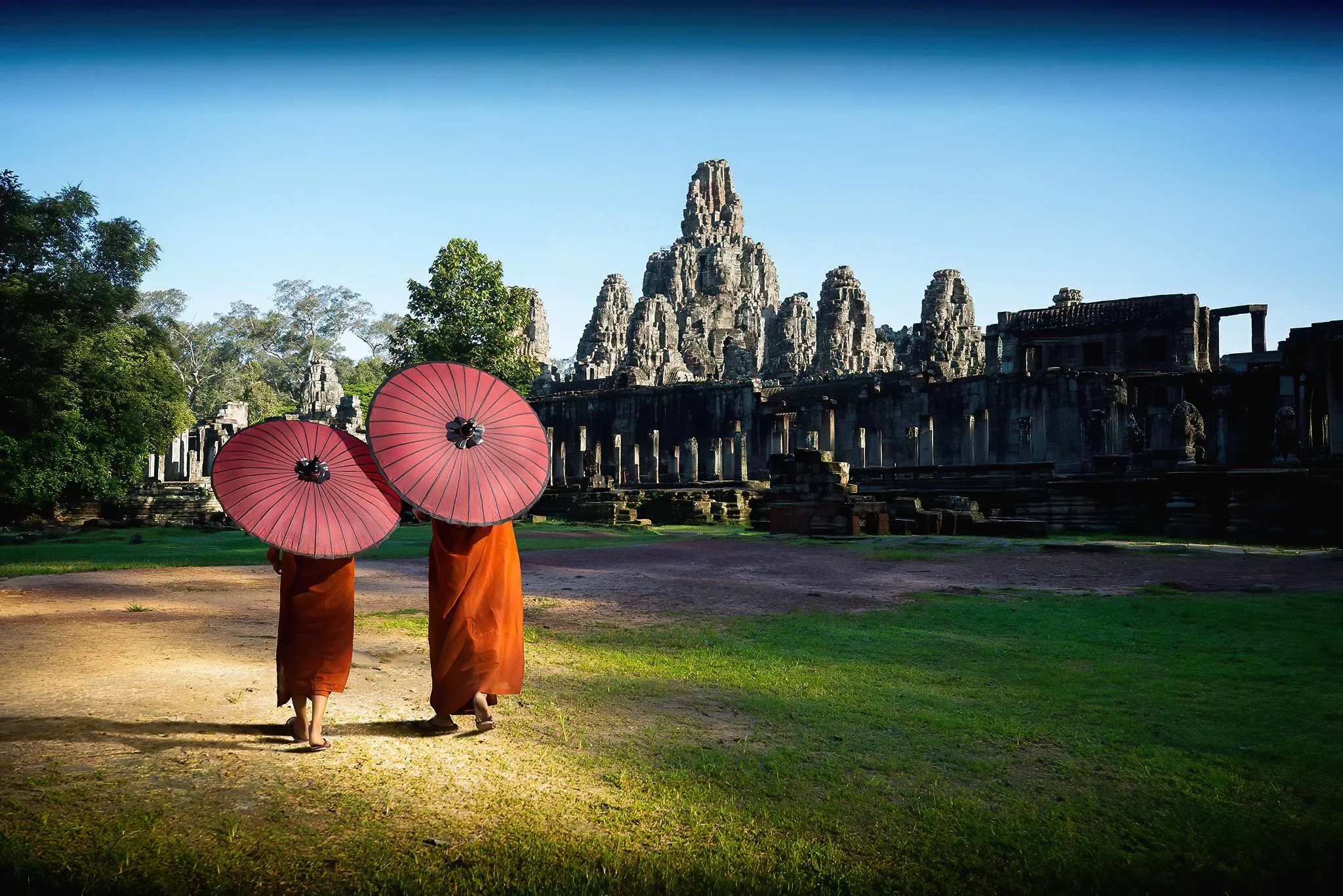 Two monks dressed in orange robes walking in front of ancient stone temple ruins, holding pink umbrellas at sunrise or sunset.