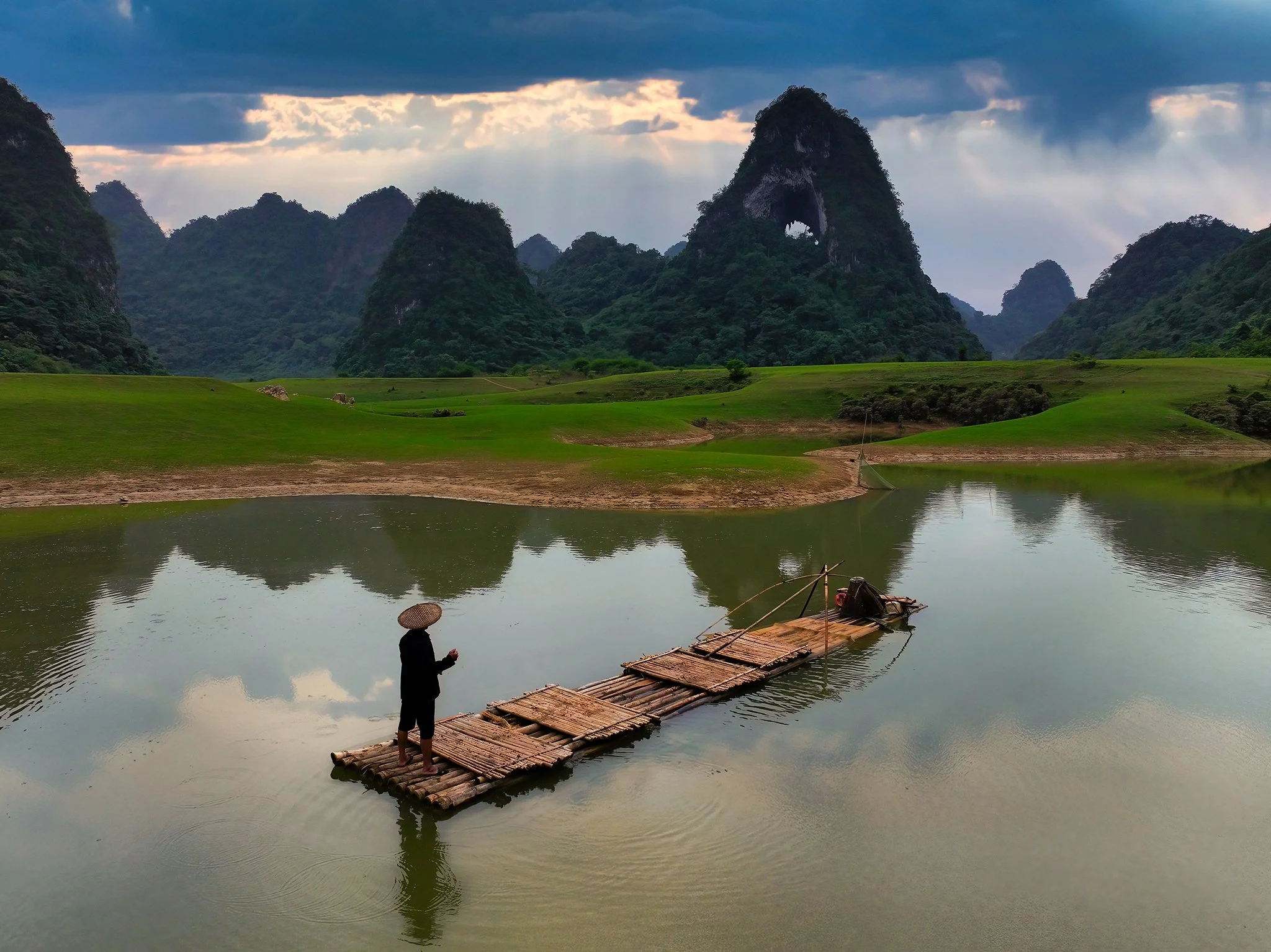 A person standing on a wooden raft on a calm river, with lush green hills and tall, steep mountains in the background. The person is wearing a hat, dark clothing, and appears to be holding a small object. The sky is partly cloudy, with sunlight breaking through clouds and illuminating the landscape.