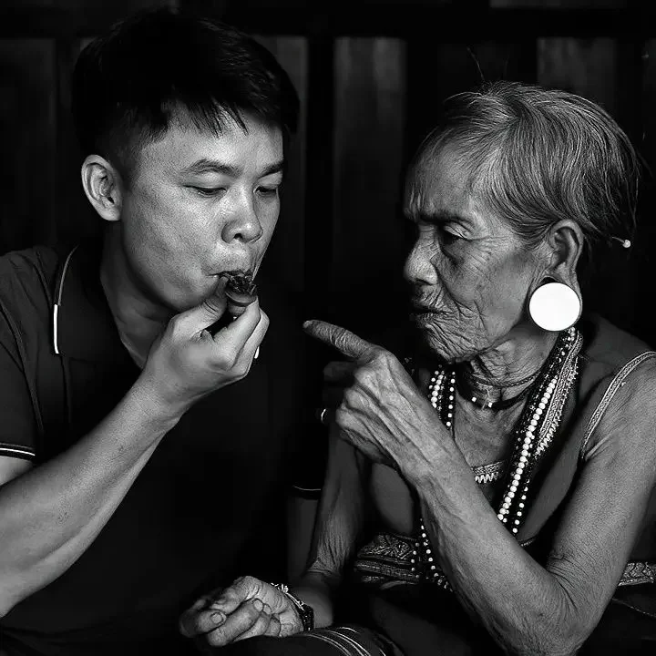 An elderly woman with earrings and necklaces pointing her finger at a young man who is eating. Both are in black and white.