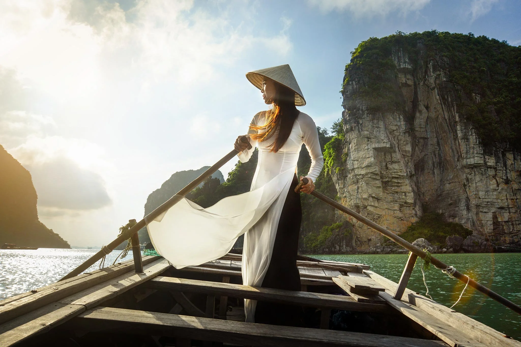 A woman in traditional Vietnamese attire and a nón lá hat standing on a wooden boat in a river, surrounded by limestone karsts.